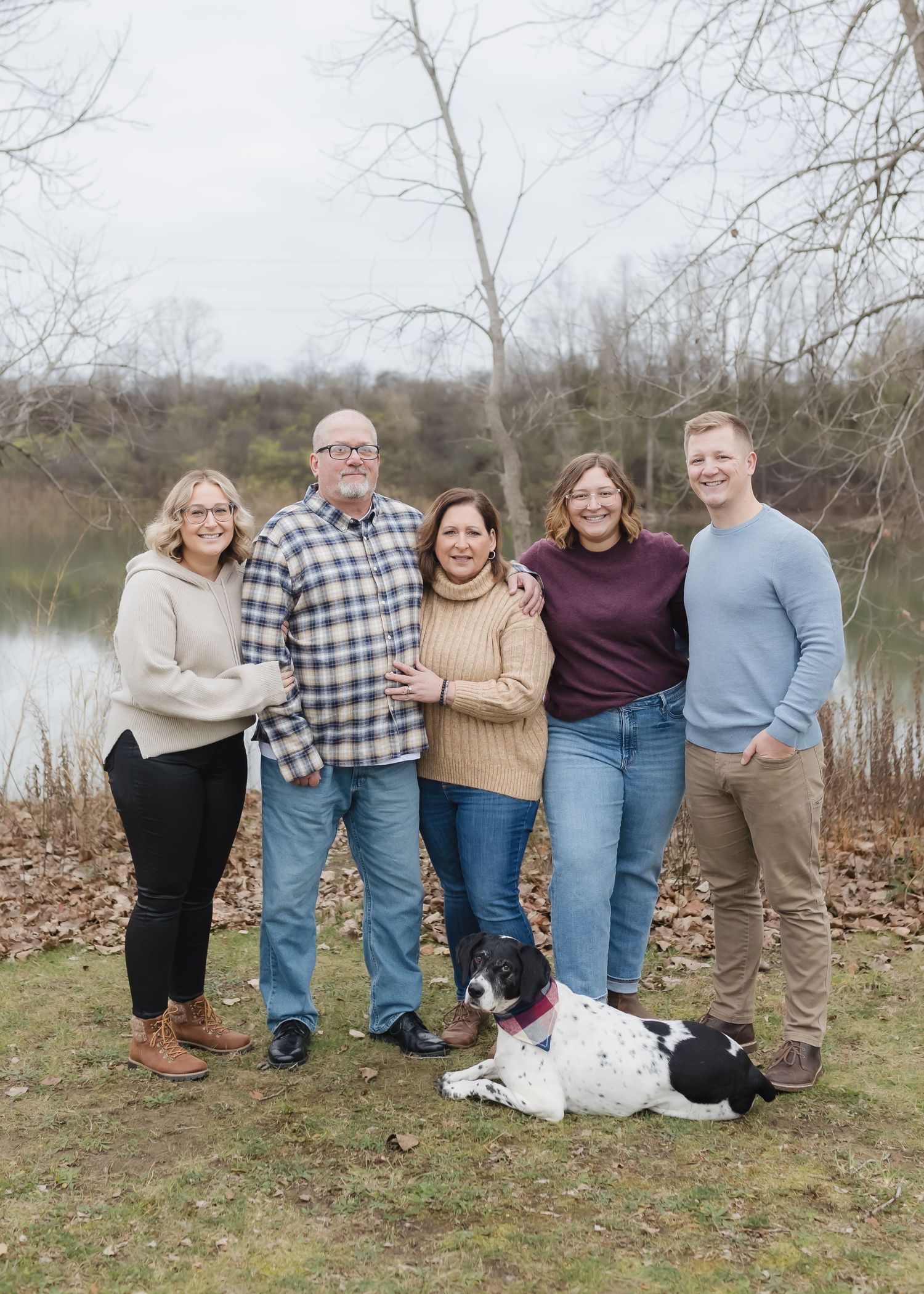 A group stands together outdoors in casual attire with a black and white dog sitting at their feet by a lake.