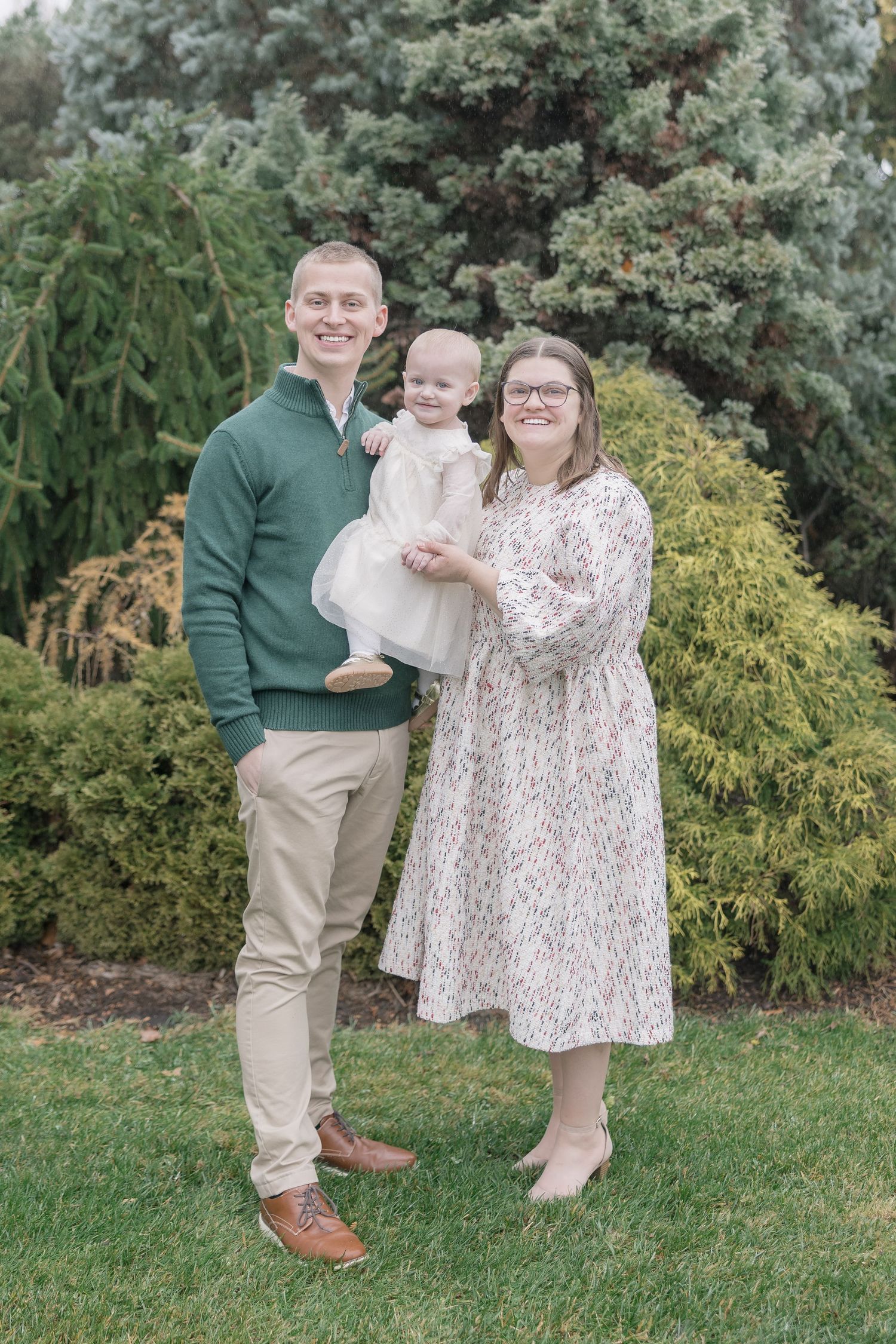 A family poses together on a grassy lawn with evergreen trees and yellow shrubs in the background.