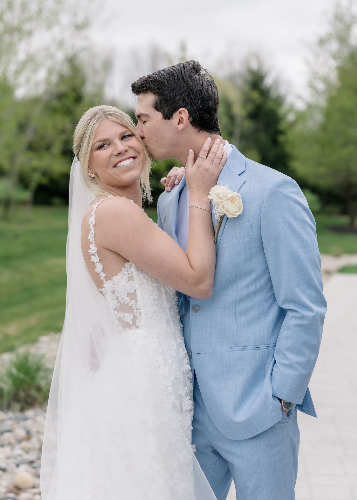 A smiling bride in a lace wedding dress and veil receives a kiss on the cheek from a groom in a light blue suit outdoors.