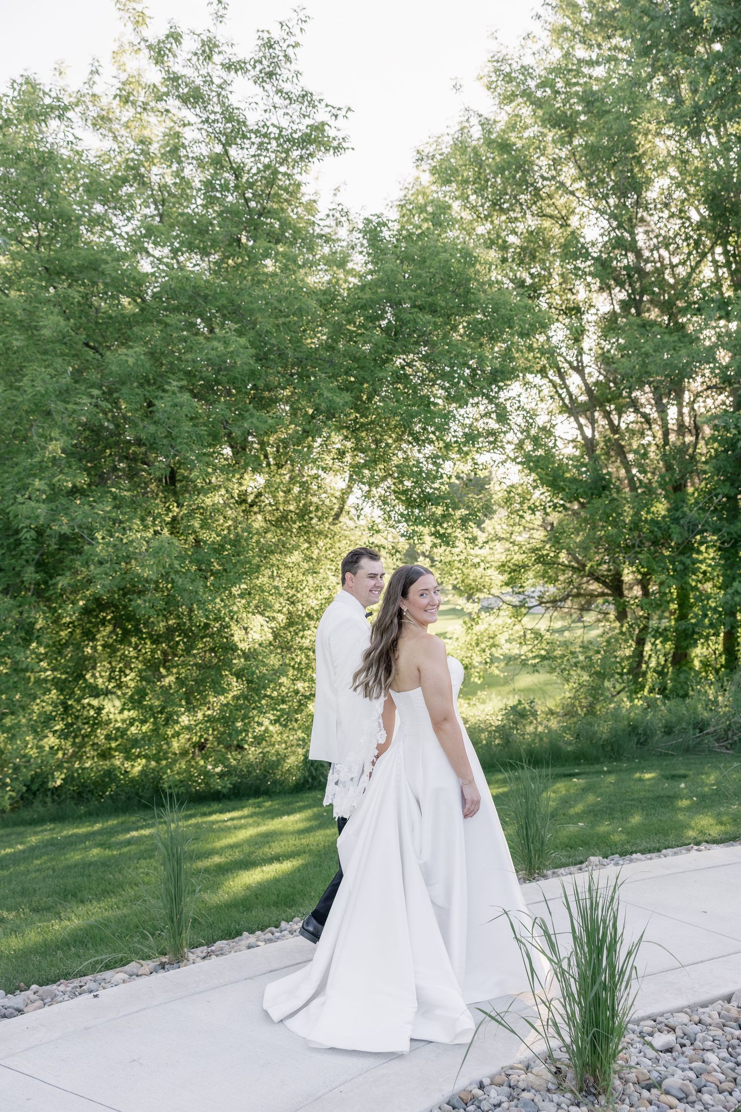 Bride and groom walk along a garden path surrounded by lush green trees on their wedding day.