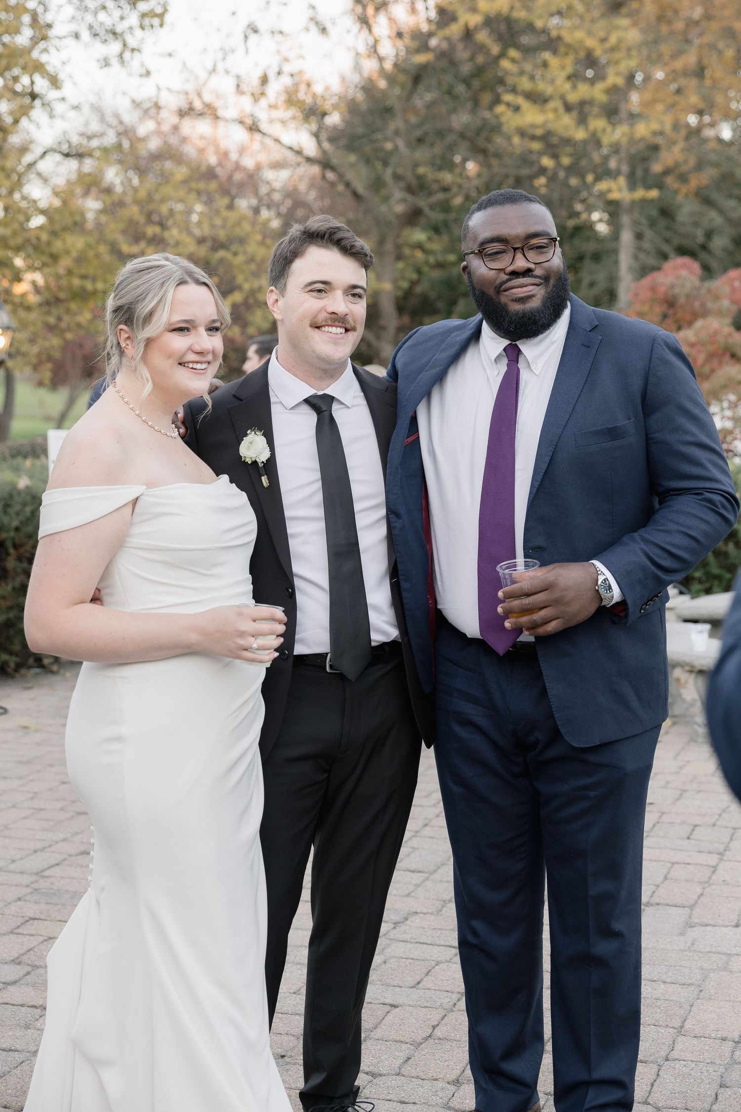Wedding party members pose together on a brick pathway during an autumn celebration.