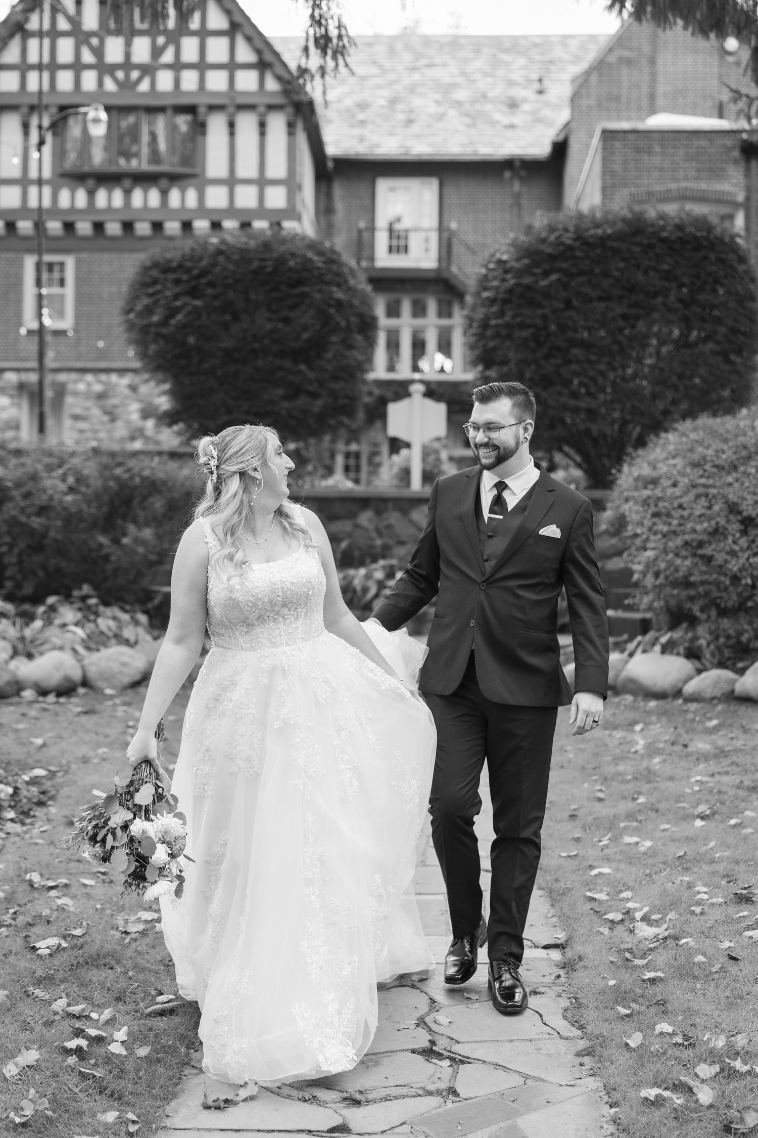 A couple walks through fallen autumn leaves in a garden setting, the bride wearing a flowing white gown.