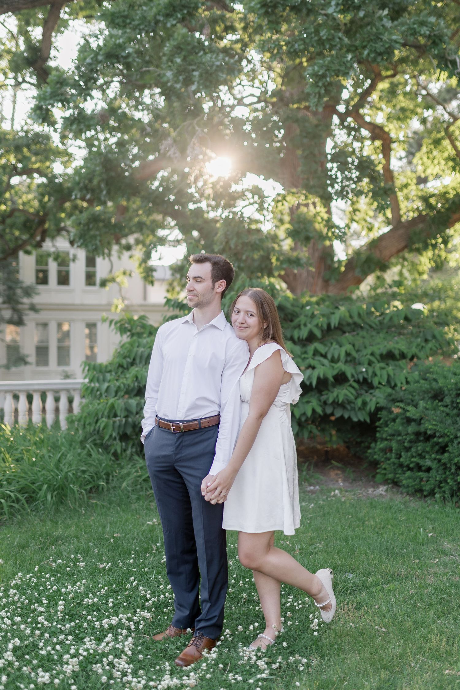 A couple poses together on a grassy lawn with sunlight streaming through trees and a classic building in the background.