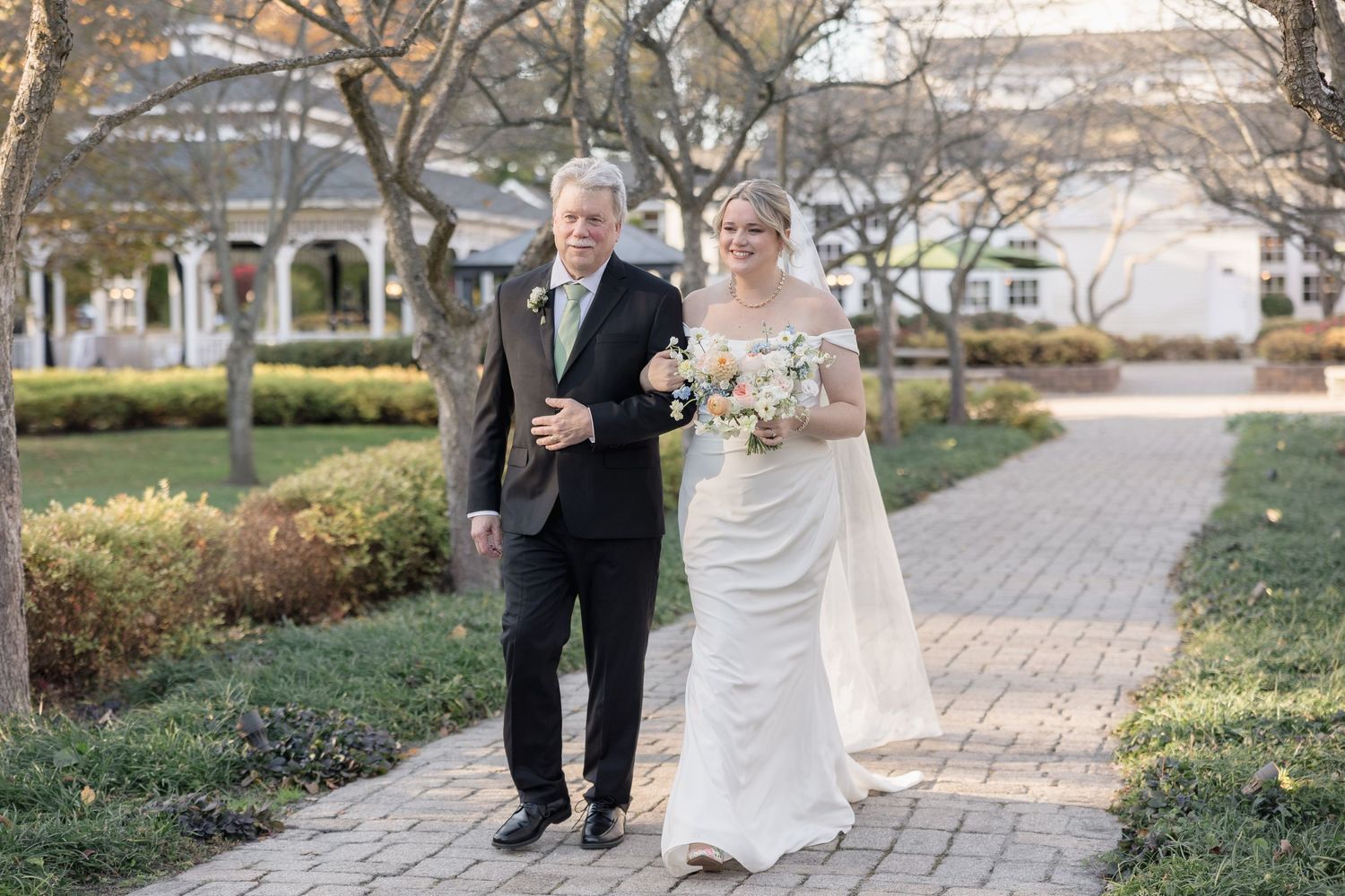 A wedding processional on a brick pathway lined with autumn trees and landscaping.