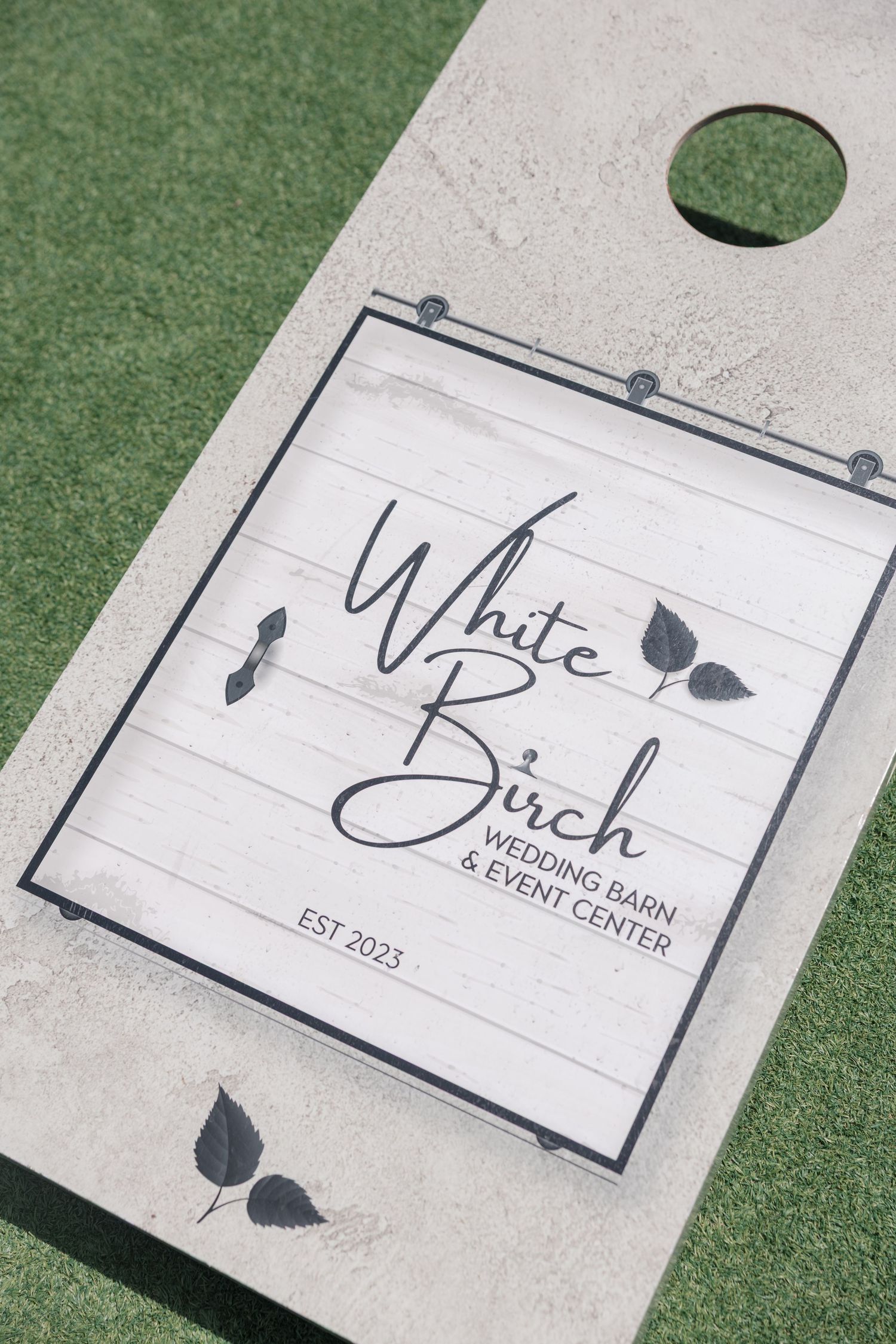 White Birch Wedding Barn and Event Center EST 2023 logo on custom cornhole board outdoors.