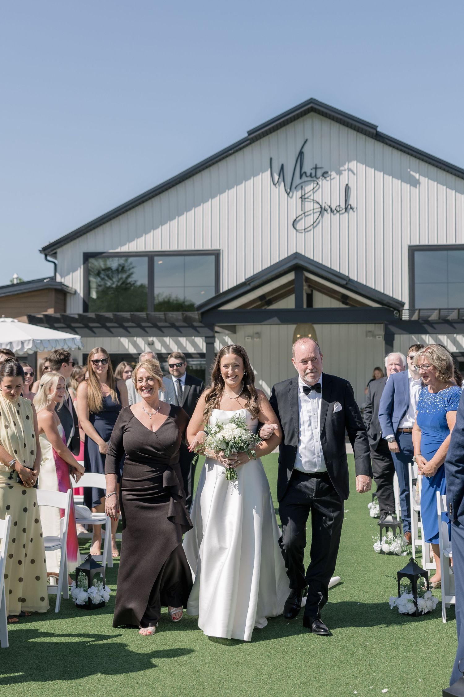 Bride walks down outdoor aisle with parents at White Birch wedding venue on sunny day.
