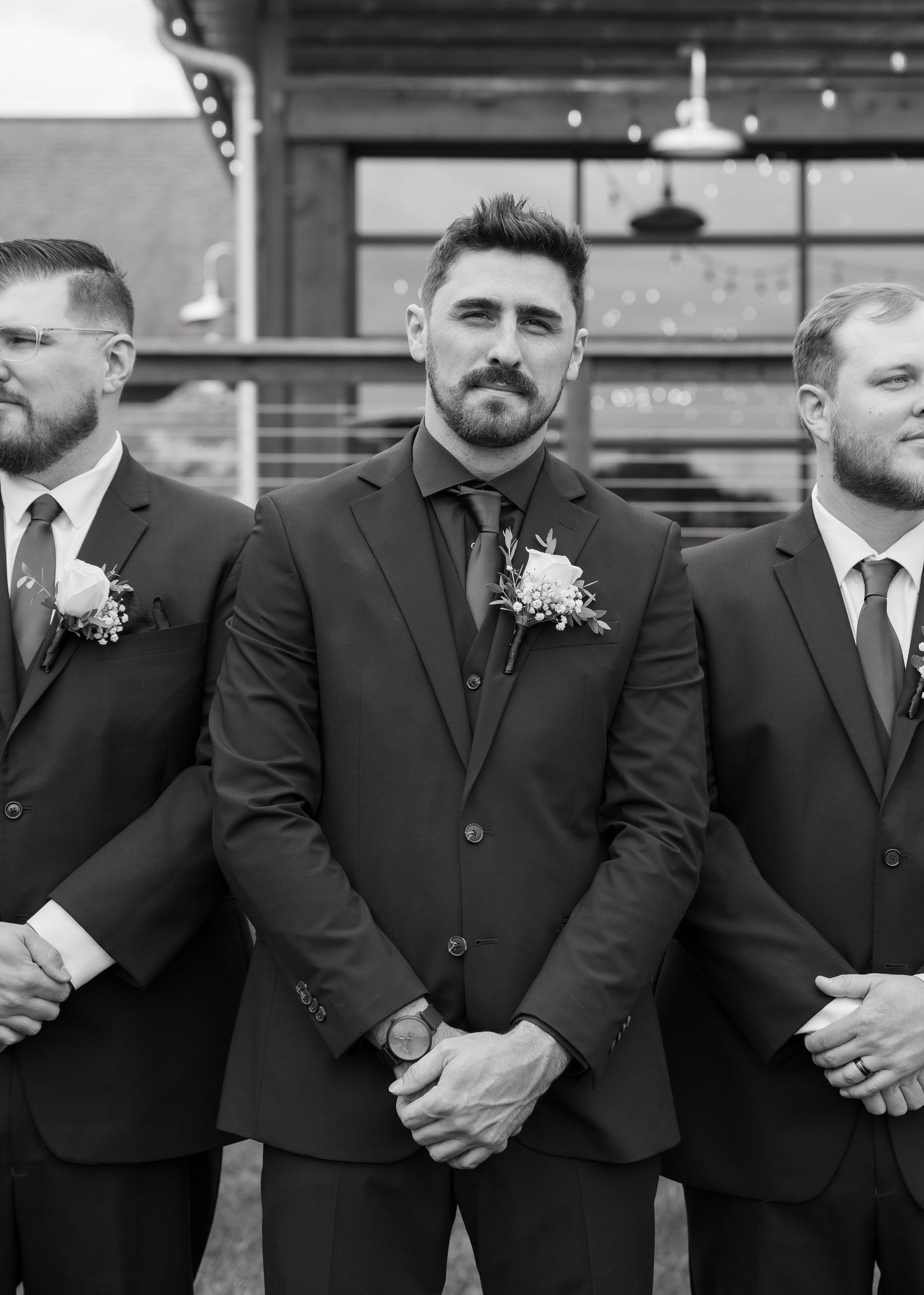A line of groomsmen in black suits with white boutonnieres stand together at a formal wedding event.