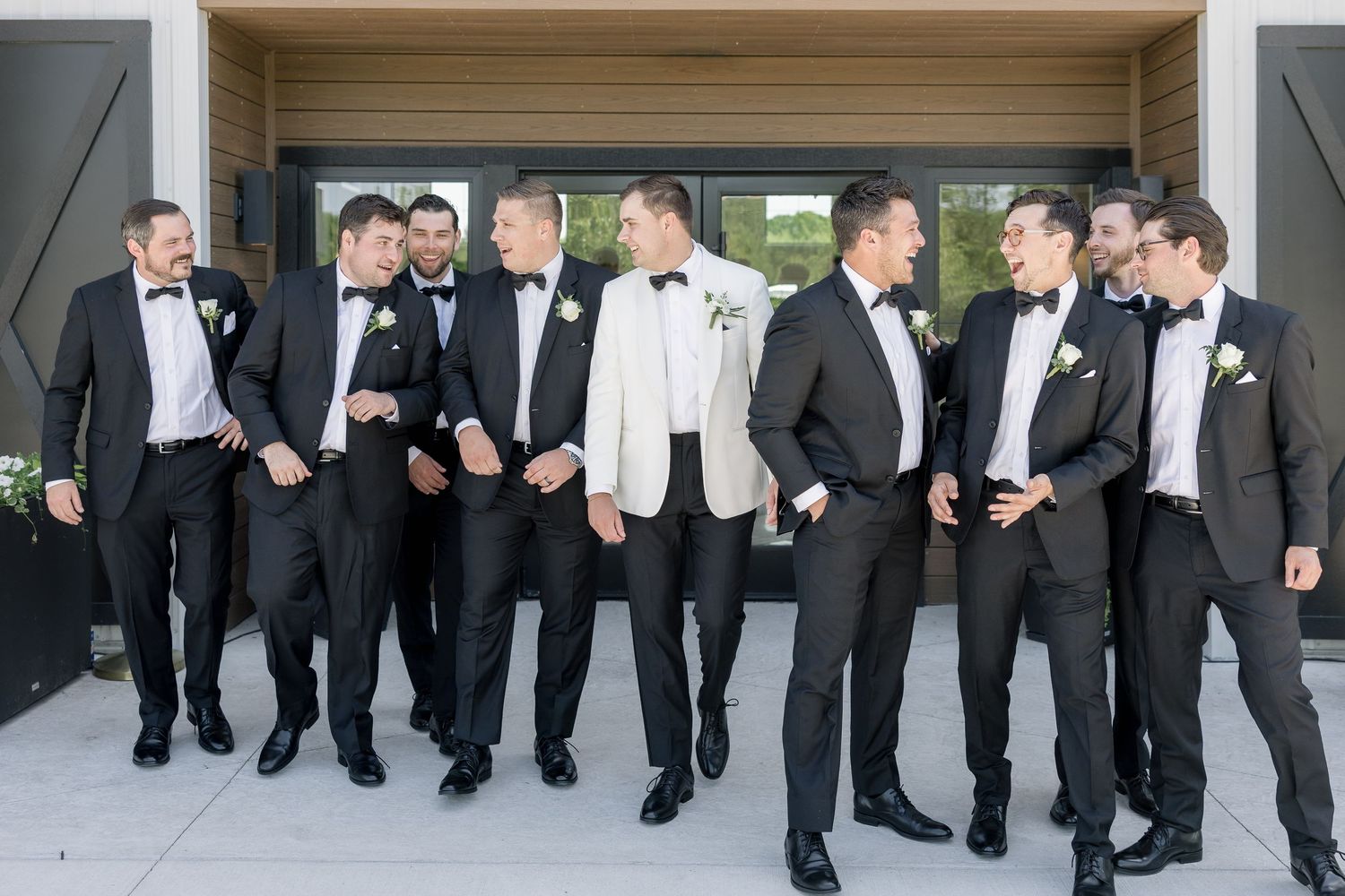 Groom in white tuxedo jacket stands with groomsmen in black tuxedos outside a modern wedding venue.