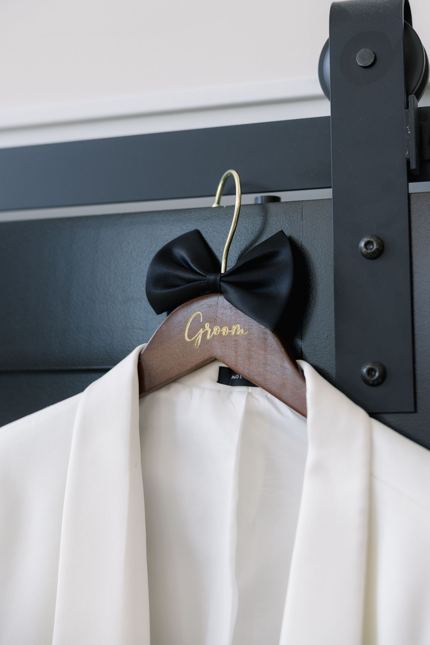 Wooden groom hanger with black bow tie on white wedding robe hung on dark barn door rail.