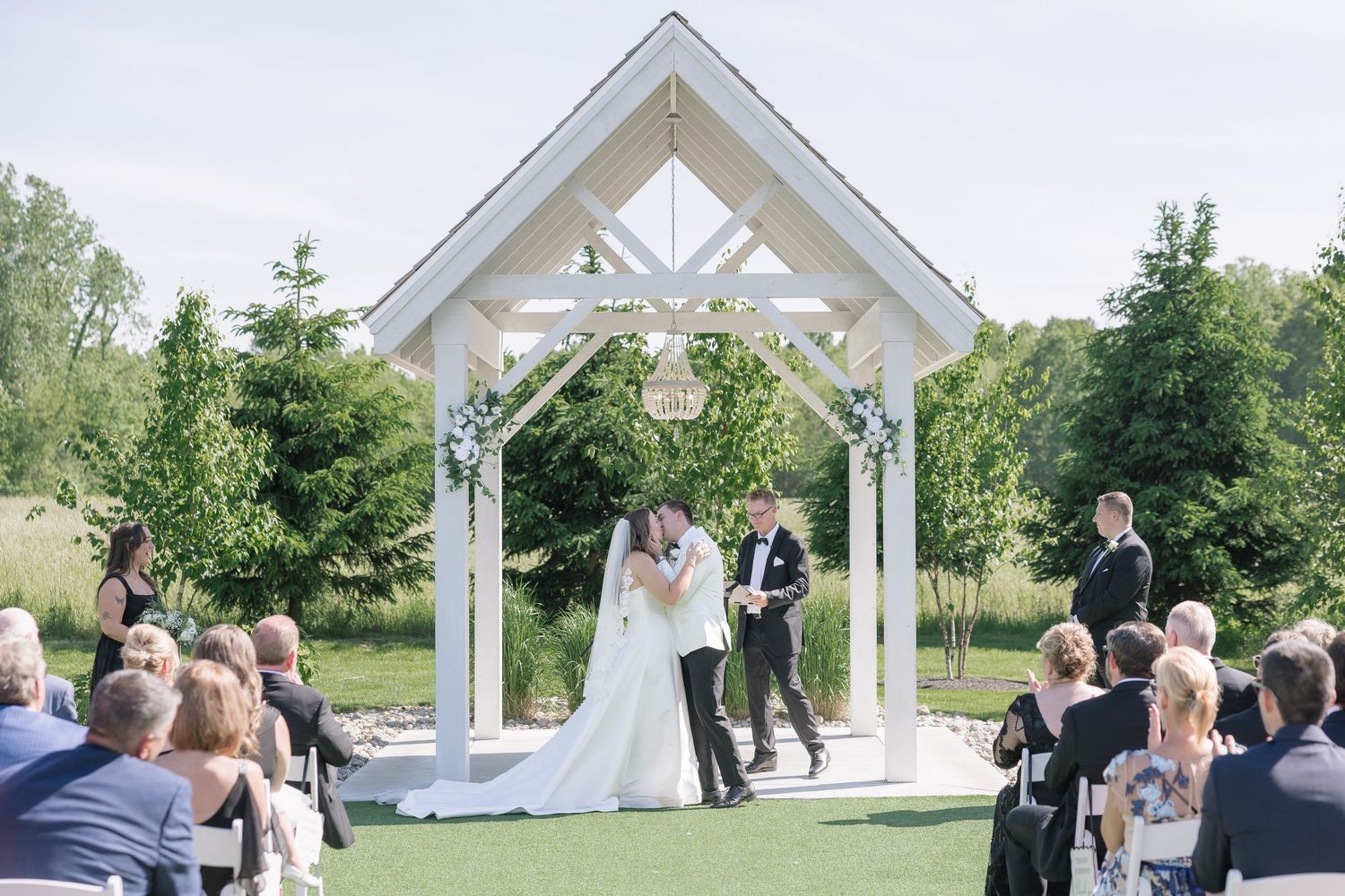 Couple shares first kiss under white wooden arch at outdoor wedding ceremony with guests seated on white chairs.