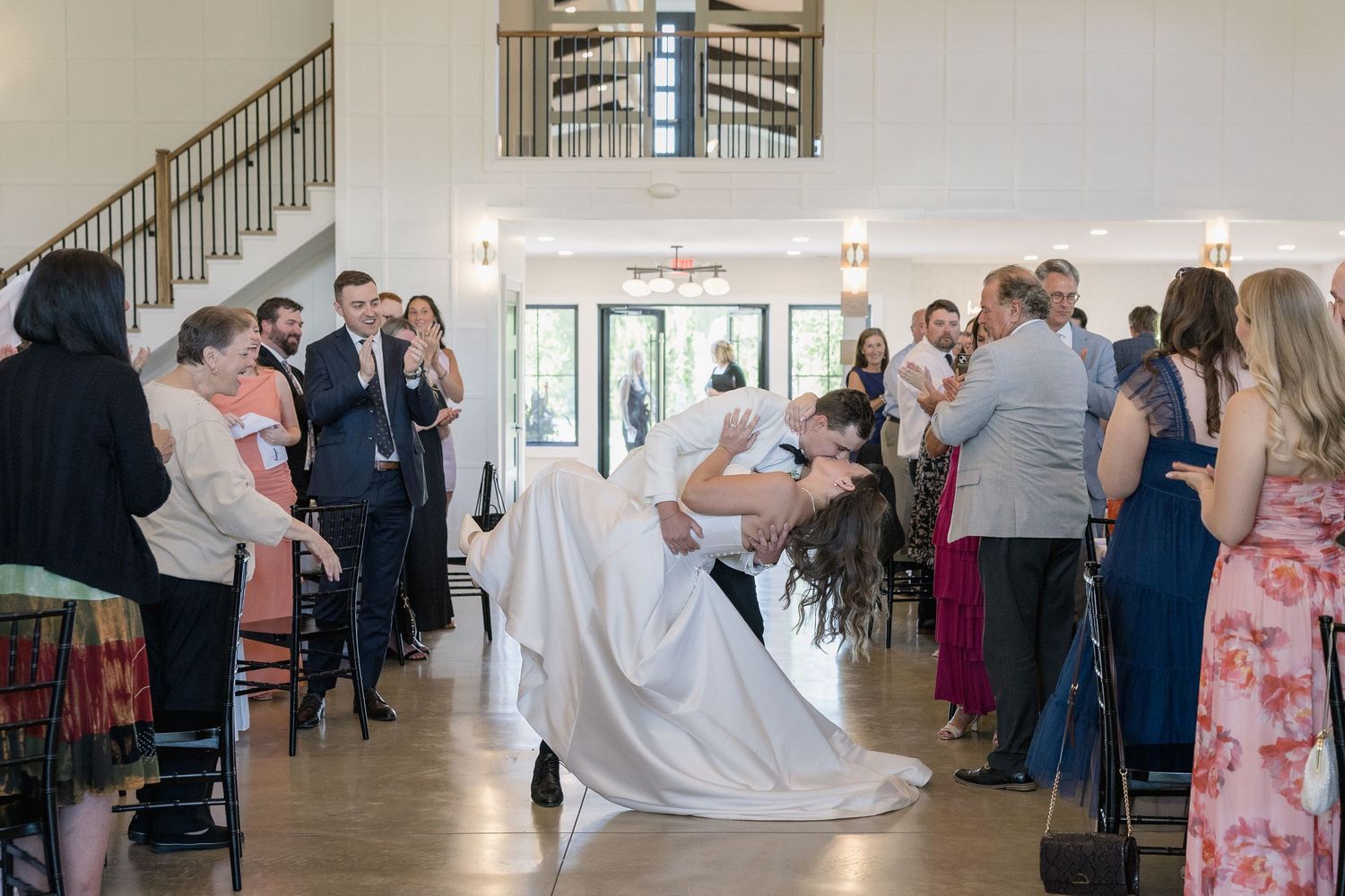 Bride and groom share a romantic dip kiss during their wedding reception while guests applaud in an elegant venue.