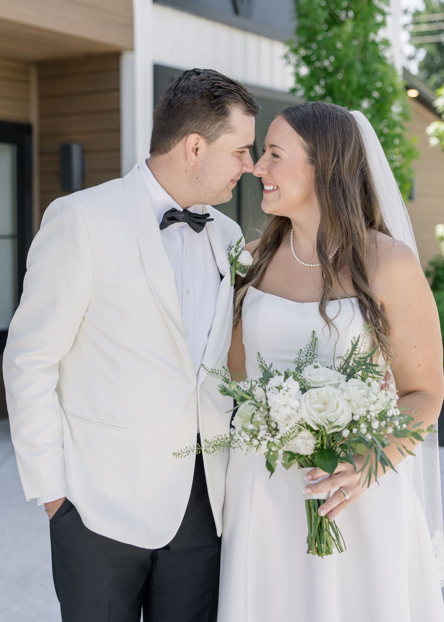 A bride and groom in wedding attire share a romantic moment, touching foreheads and smiling outdoors.