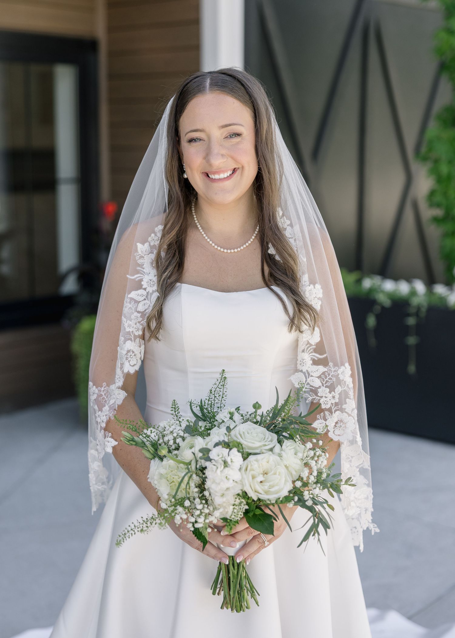 Smiling bride in a white gown and lace veil holding a bouquet of white roses and greenery on her wedding day.