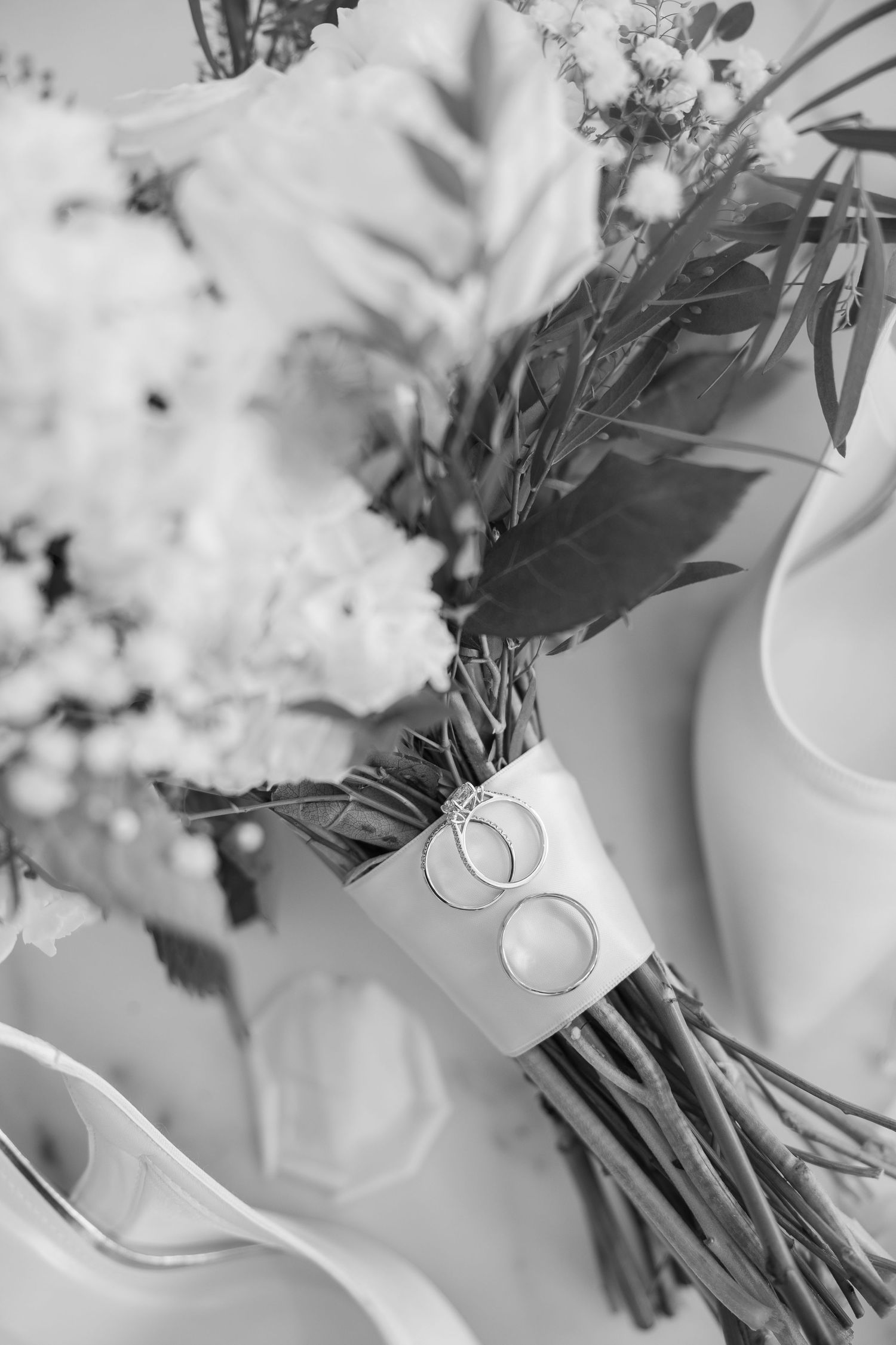 Black and white wedding bouquet with rings tied to stems, beside bridal heels and floral arrangement.