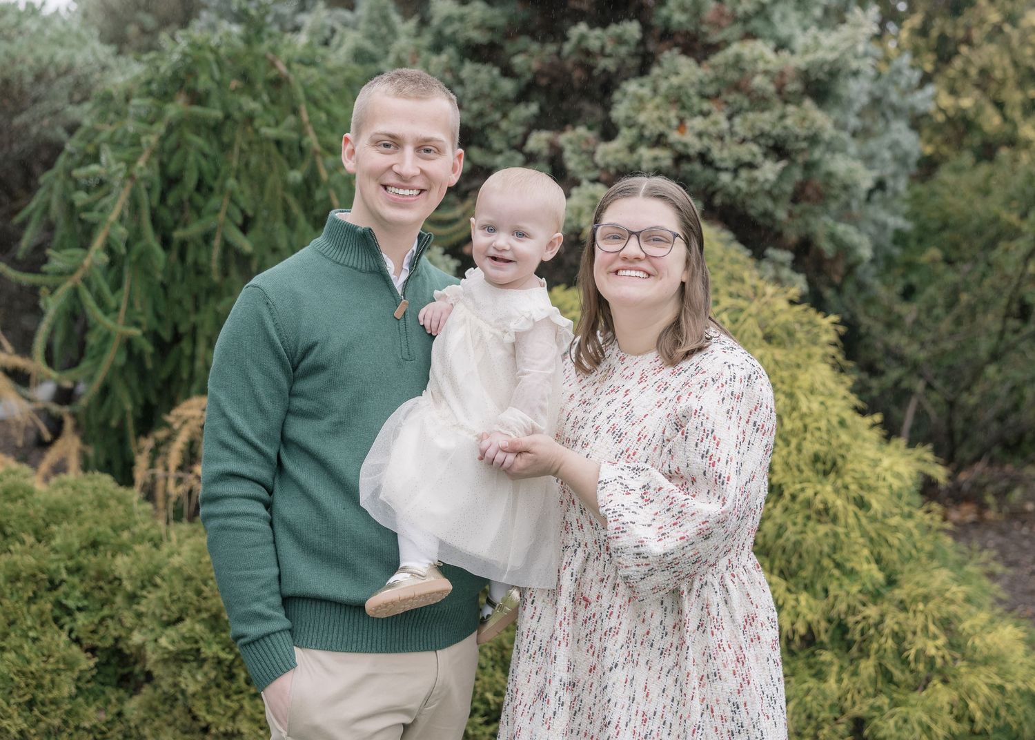 A smiling family of three poses outdoors, with a man in green holding a baby girl in white dress beside a woman in floral dress.