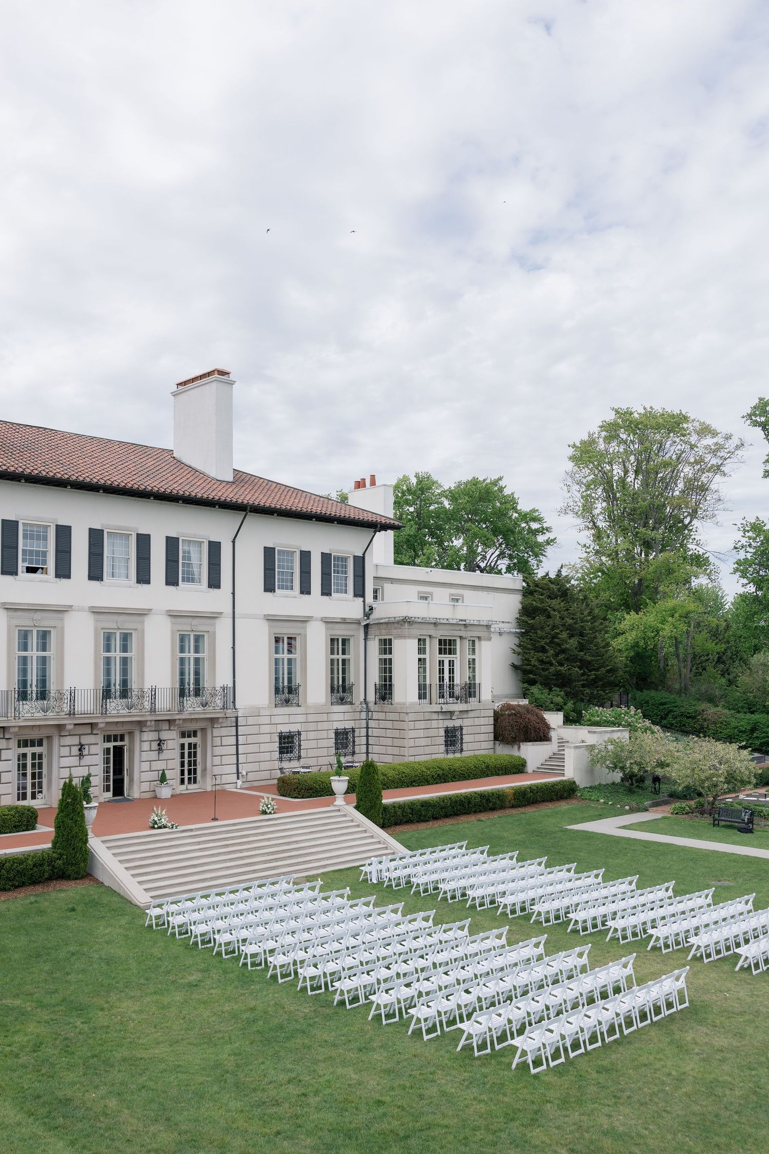 White chairs arranged in rows on a manicured lawn outside an elegant mansion for an outdoor wedding ceremony.