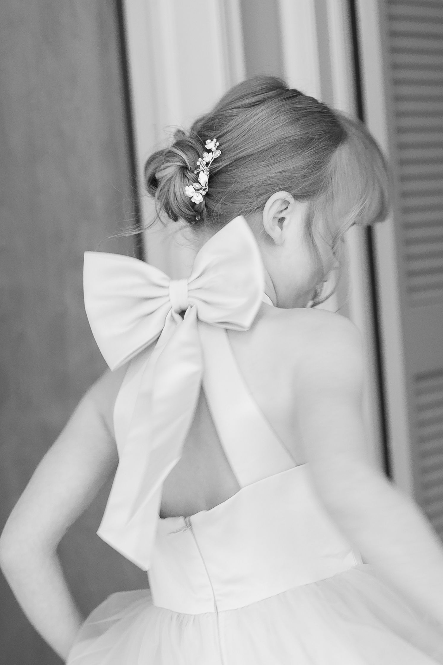 Back view of a flower girl's dress featuring a large white bow and floral hair accessory.