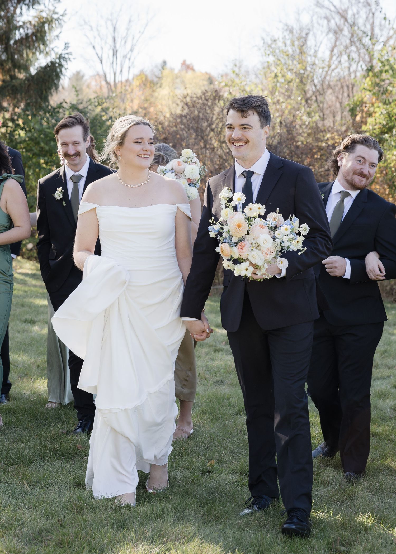 Wedding party walks and laughs together on a grassy lawn on a sunny autumn day with the bridal bouquet in view.