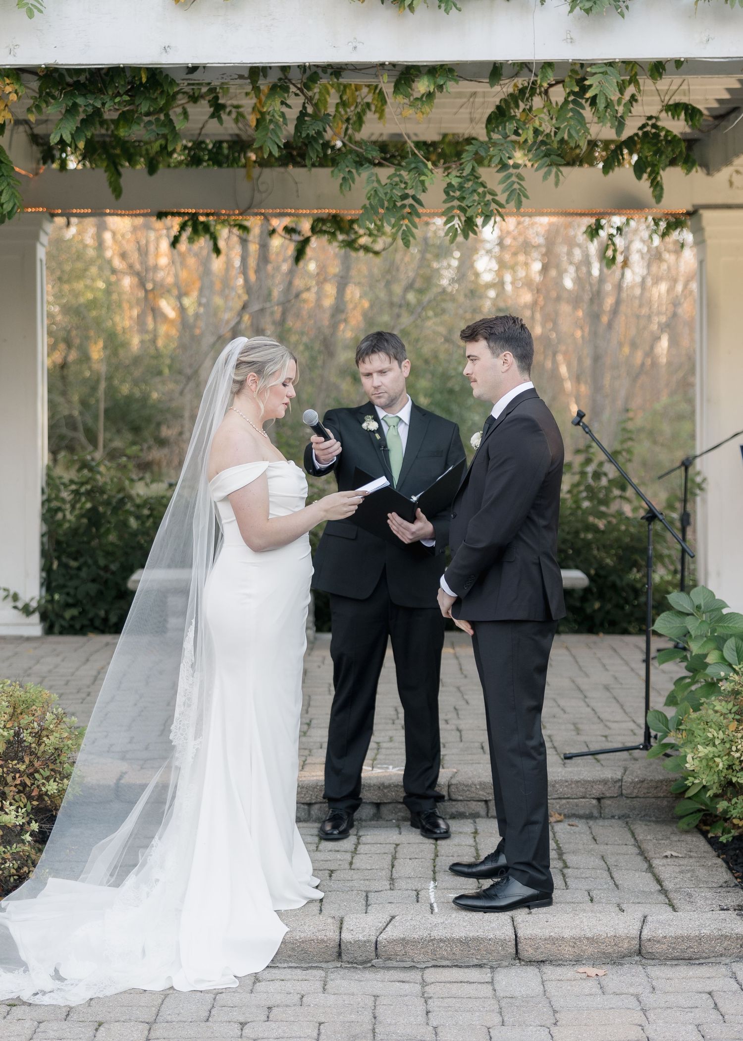 Wedding ceremony unfolds under pergola with falling autumn leaves in background.