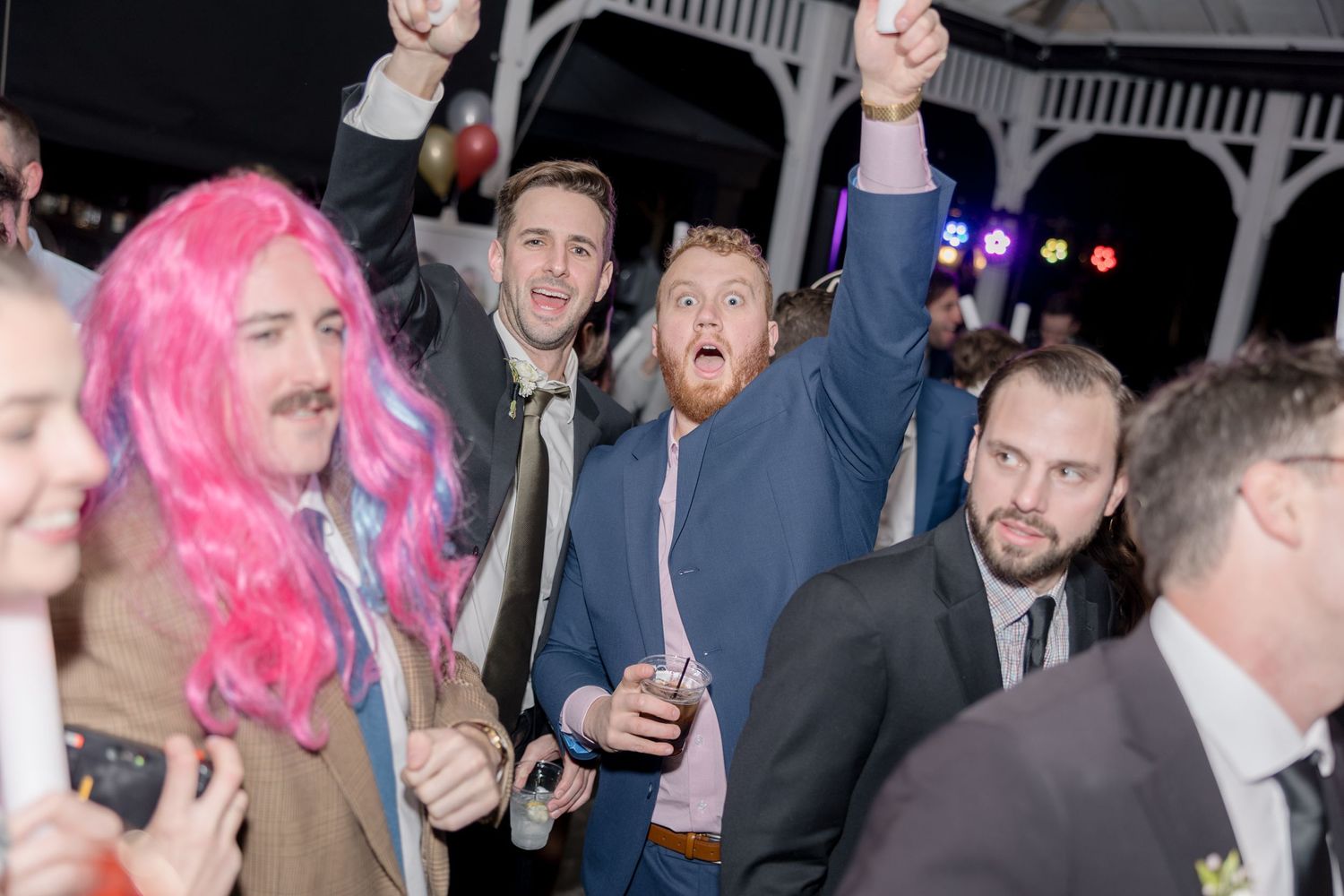 Energetic wedding reception crowd cheering with one attendee wearing a bright pink wig.