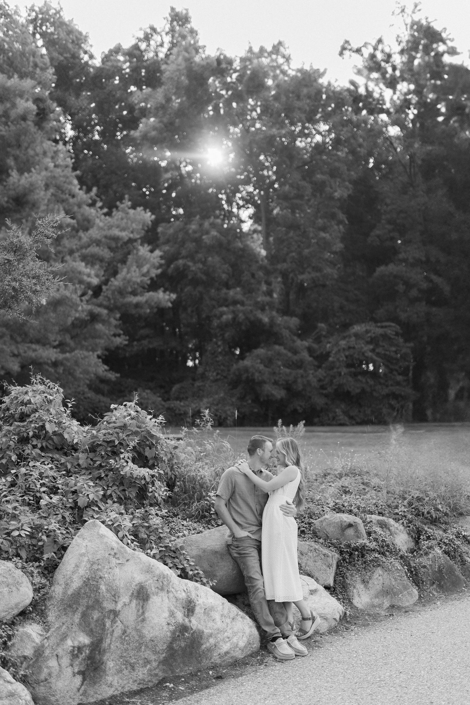 A romantic black and white photo of a couple embracing by large rocks in a park with sunlight filtering through trees.