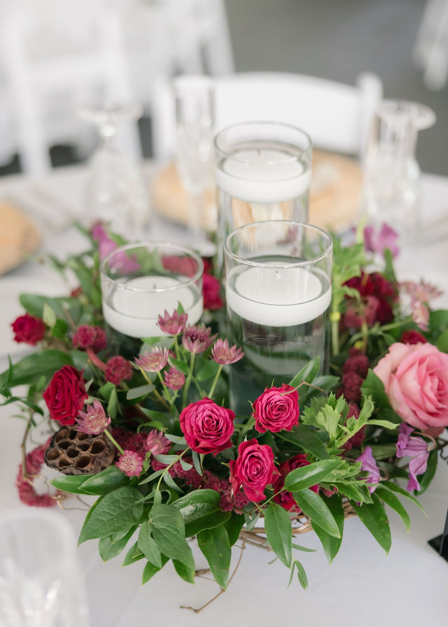 A wedding table centerpiece features pink roses, greenery and white candles in glass holders arranged in a circular pattern.