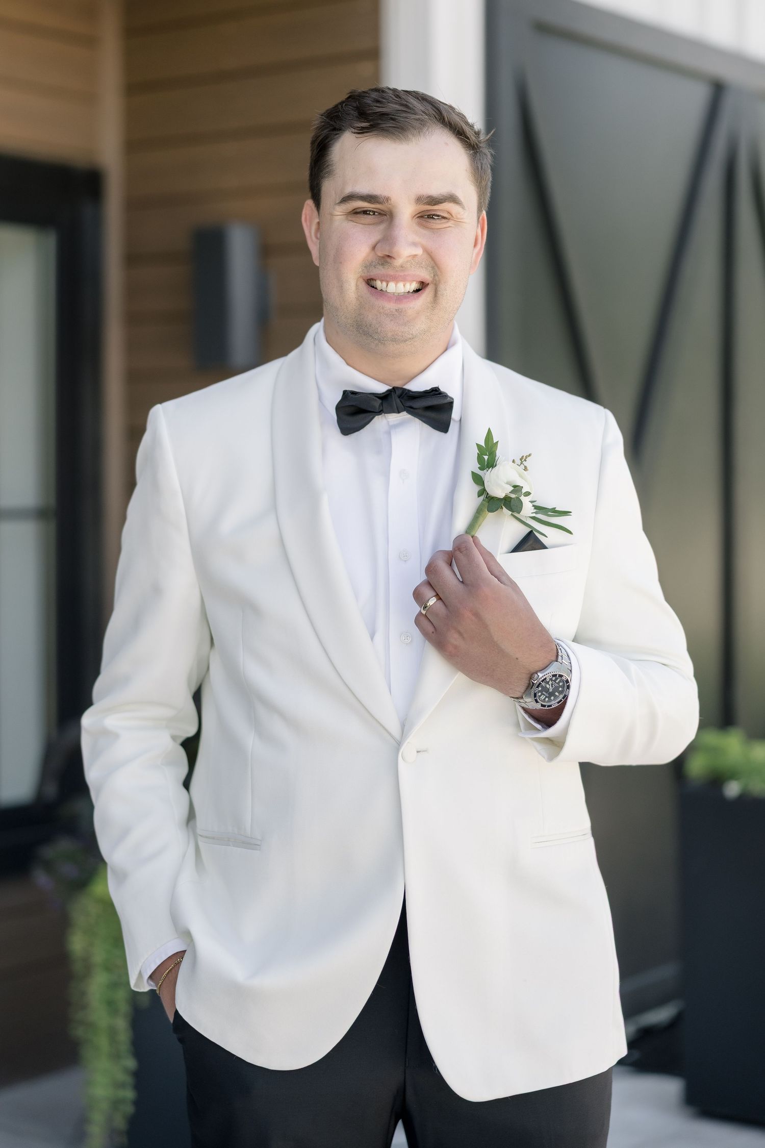 Smiling groom in white tuxedo jacket with black bow tie and floral boutonniere poses outdoors on wedding day.