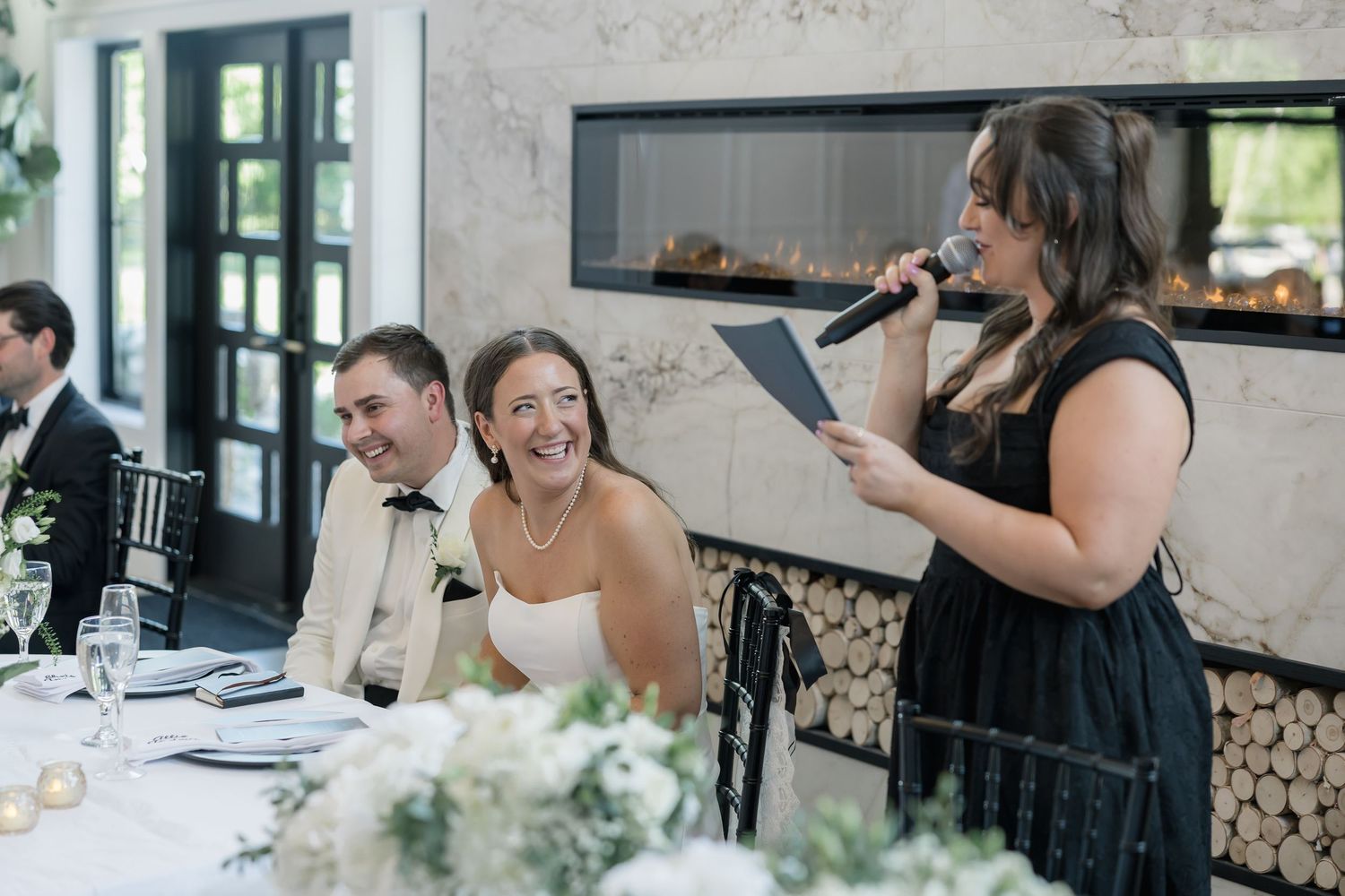Bride and groom laugh during wedding reception speech given by woman in black dress at elegant venue.