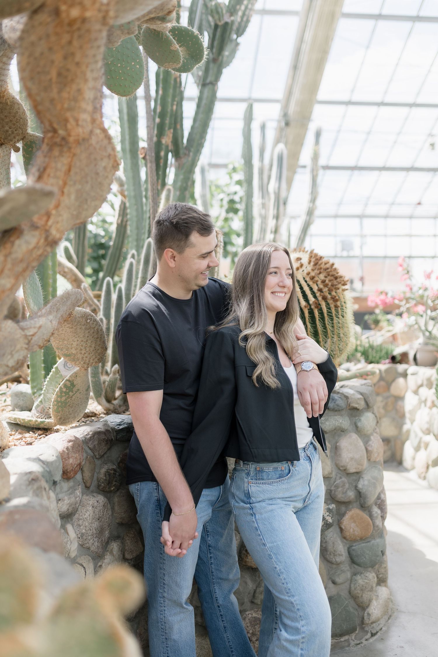A couple shares a romantic moment in a greenhouse filled with cacti and natural light.