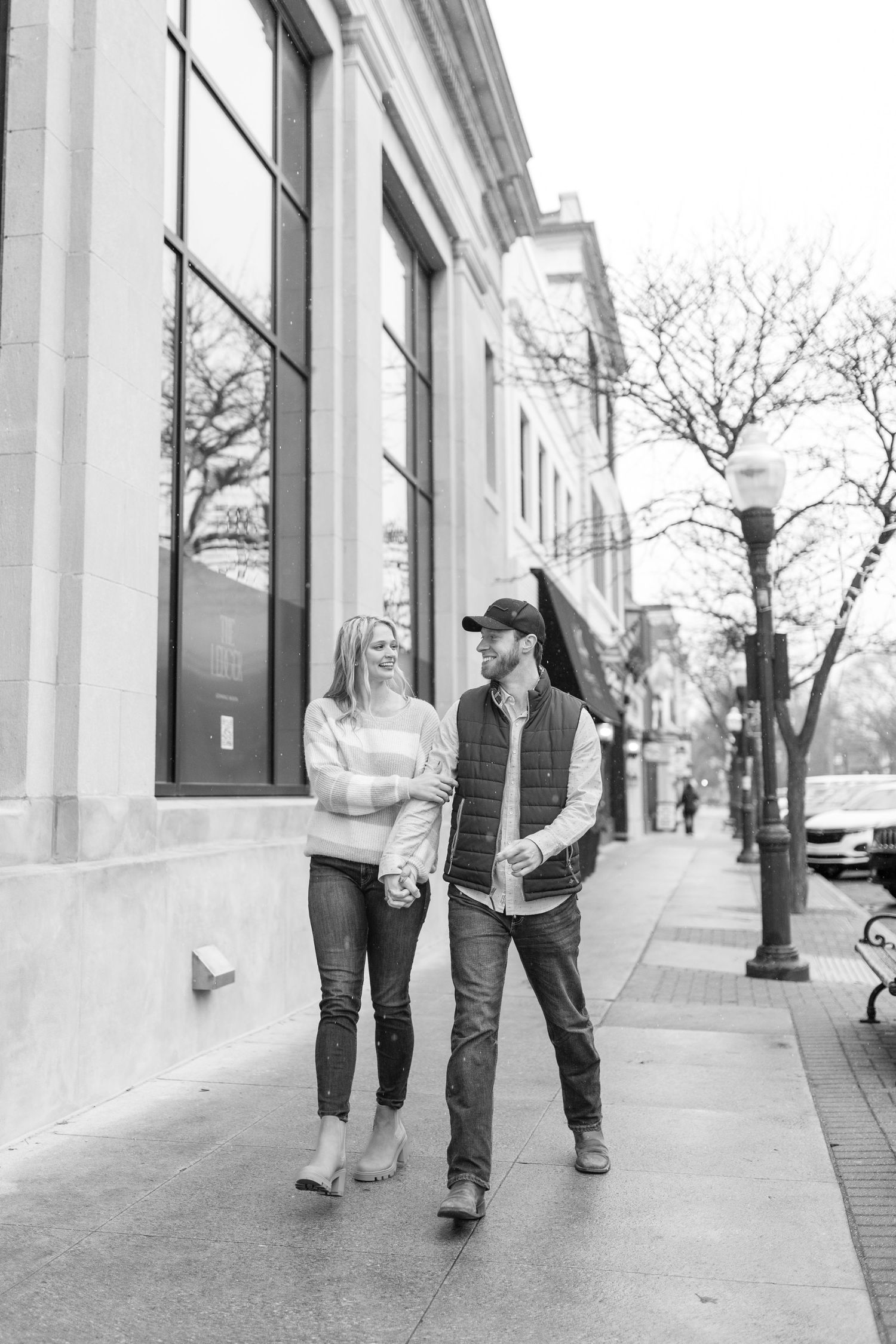 A couple strolls together along a city sidewalk in black and white photography.