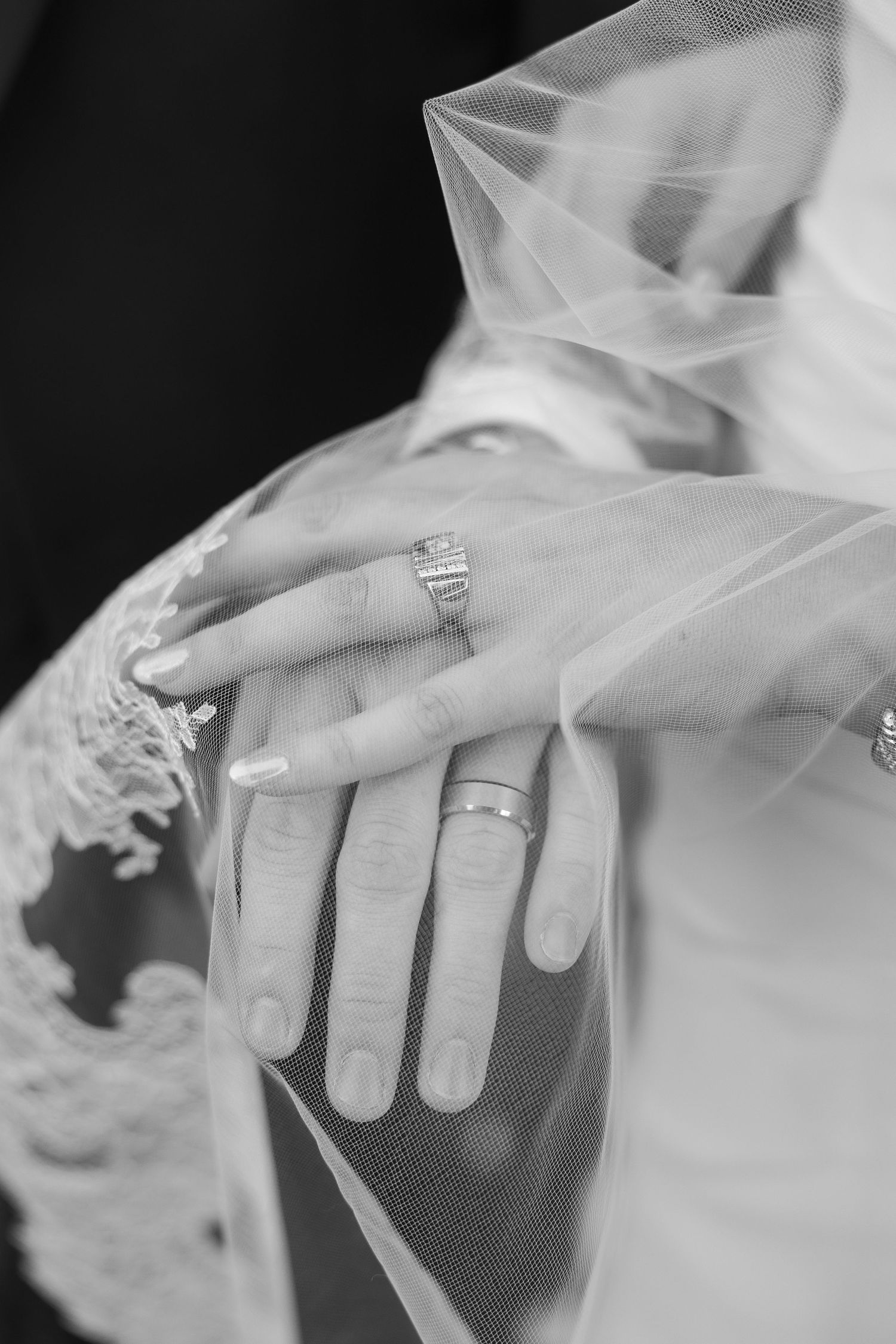 Close up black and white photo of wedding rings and manicured hands under delicate veil.