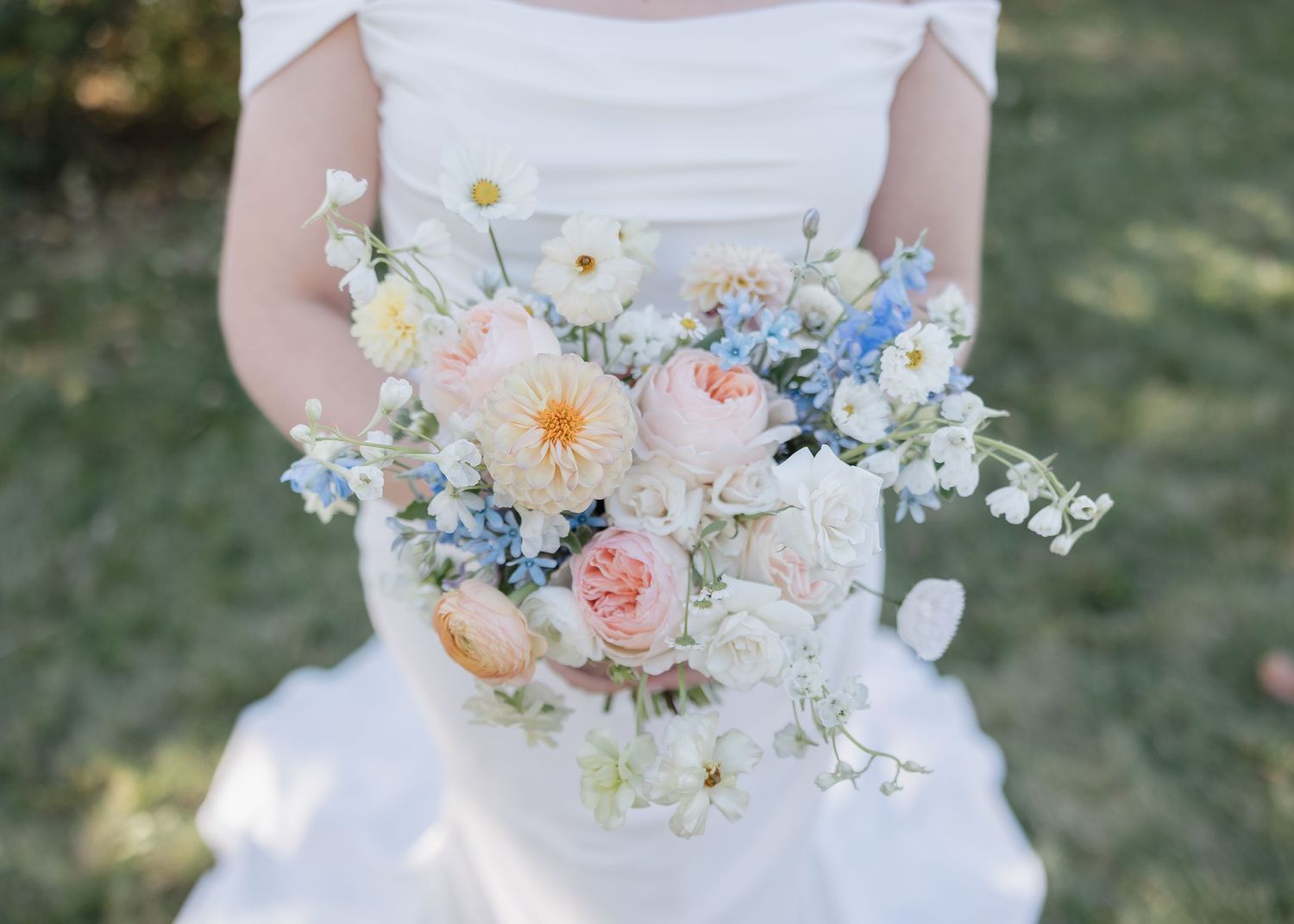 A delicate pastel wedding bouquet with blush roses, blue delphiniums, white daisies and soft peach blooms against a white dress.