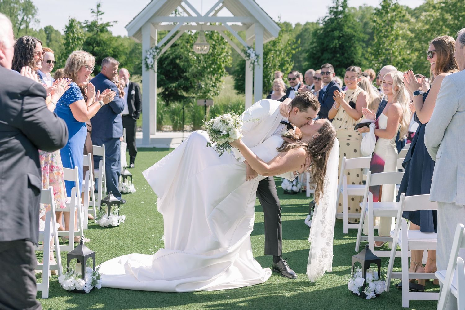 Bride and groom share a romantic dip kiss at outdoor wedding ceremony aisle surrounded by applauding guests.