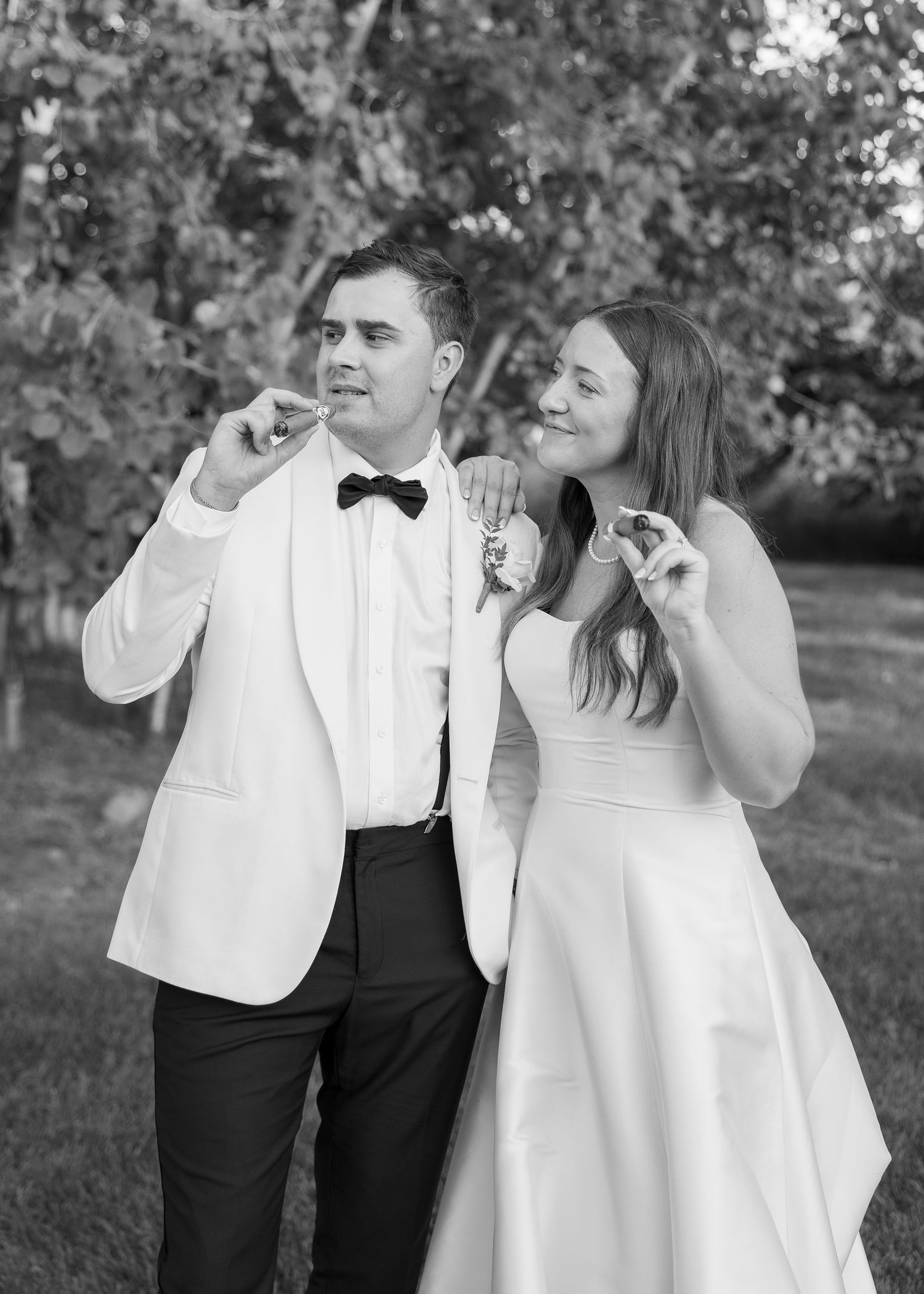 Black and white photo of a bride and groom in wedding attire smiling outdoors, holding small treats near their faces.