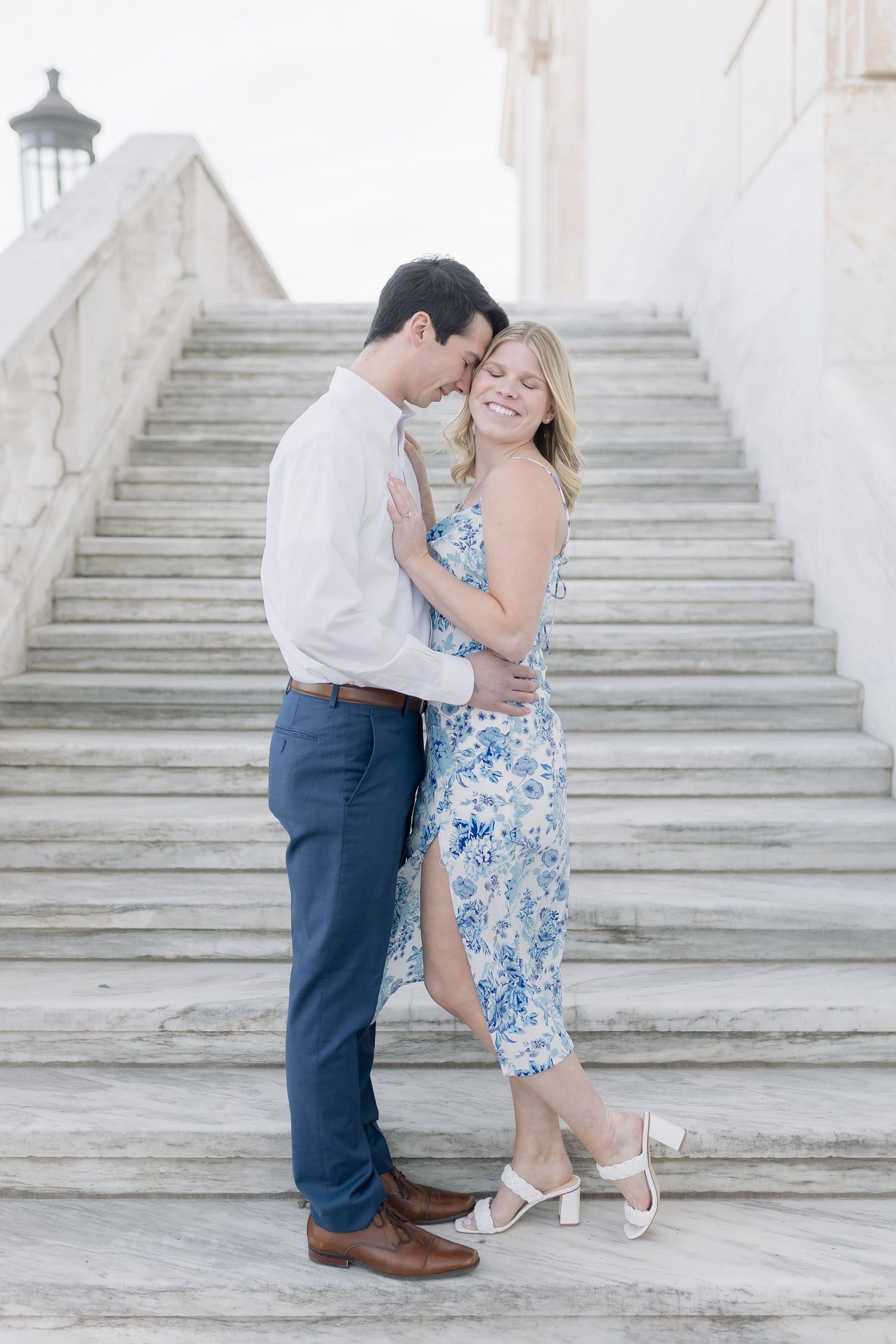 A couple poses romantically on white stone steps wearing coordinated blue and white outfits.