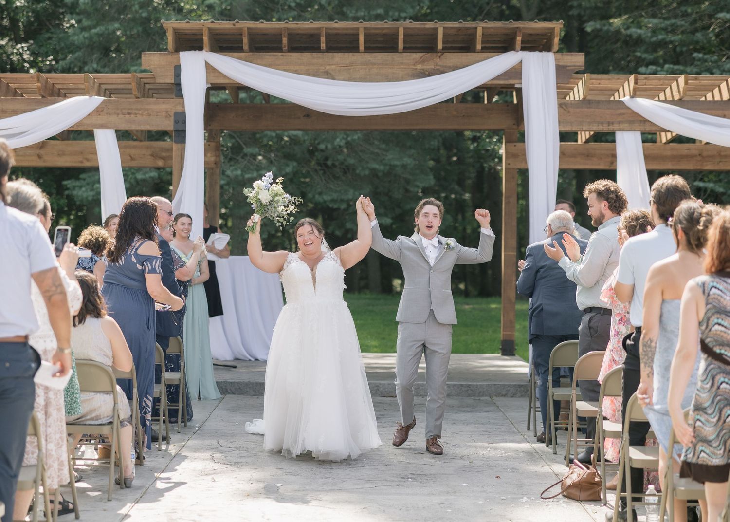 A wedding party celebrates under an outdoor pavilion decorated with white drapes and wooden beams.