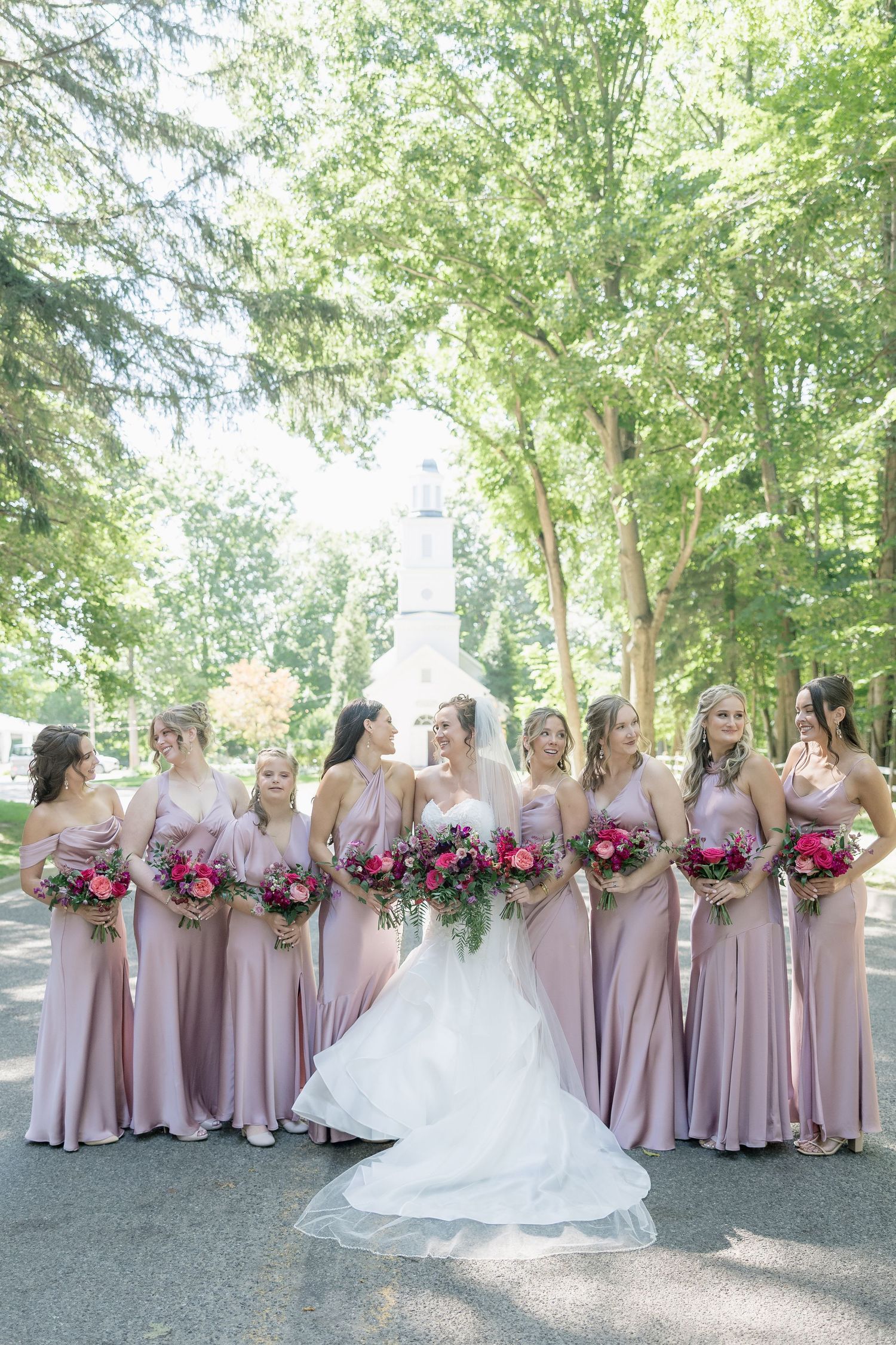 Bridal party in blush pink dresses holding vibrant pink and white bouquets pose together on a tree-lined path.