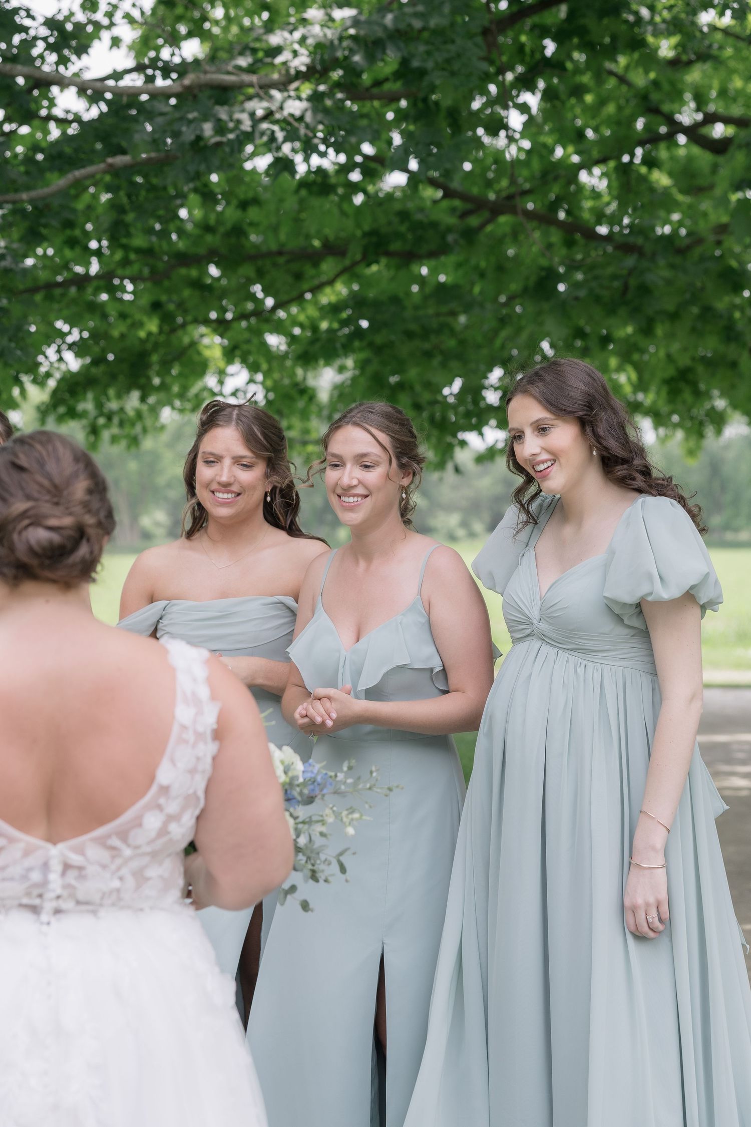Bridesmaids in soft sage green dresses share a joyful moment with the bride during an outdoor wedding celebration.