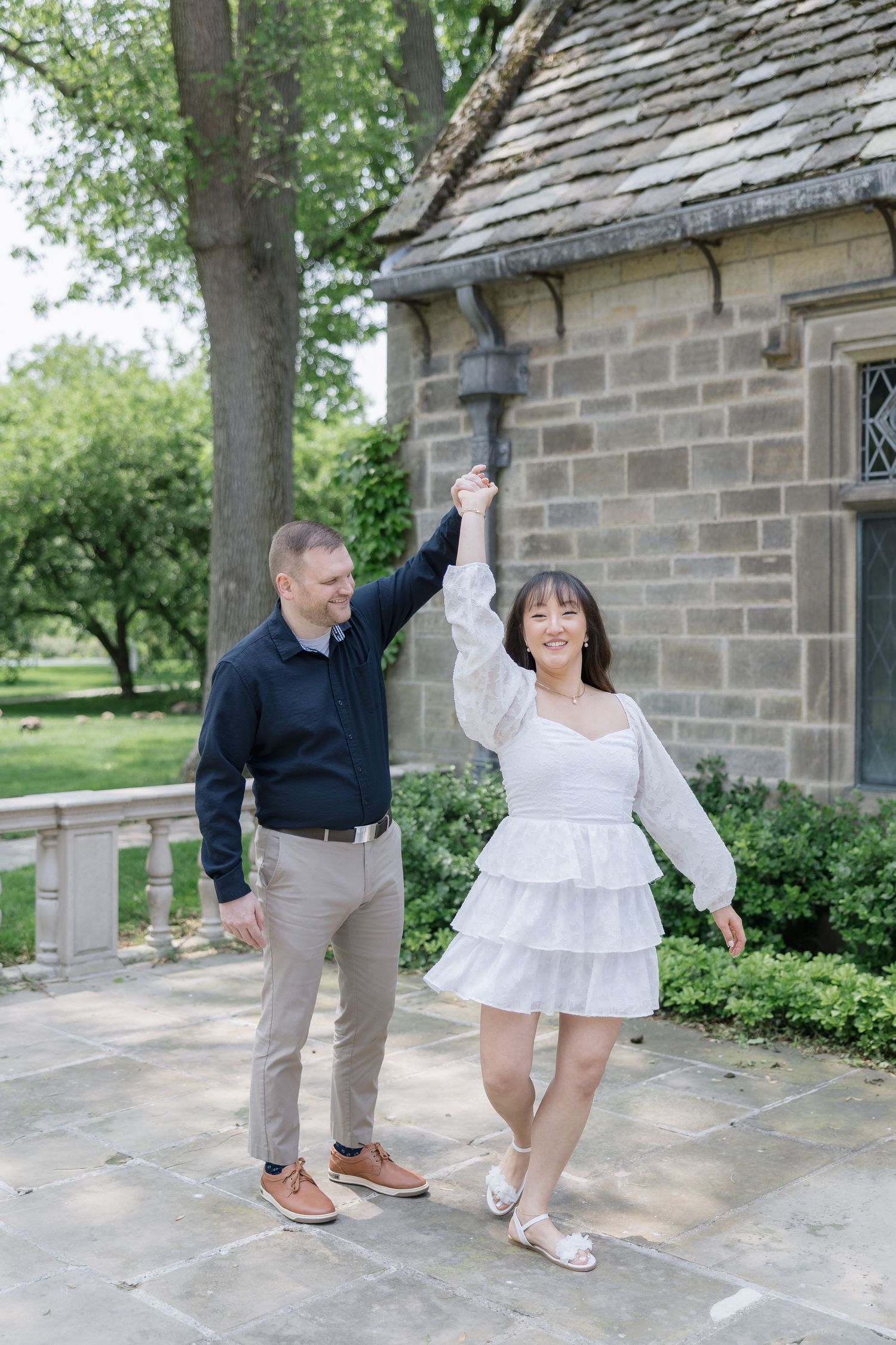 A couple dances joyfully together on a stone patio outside a historic building with greenery in the background.