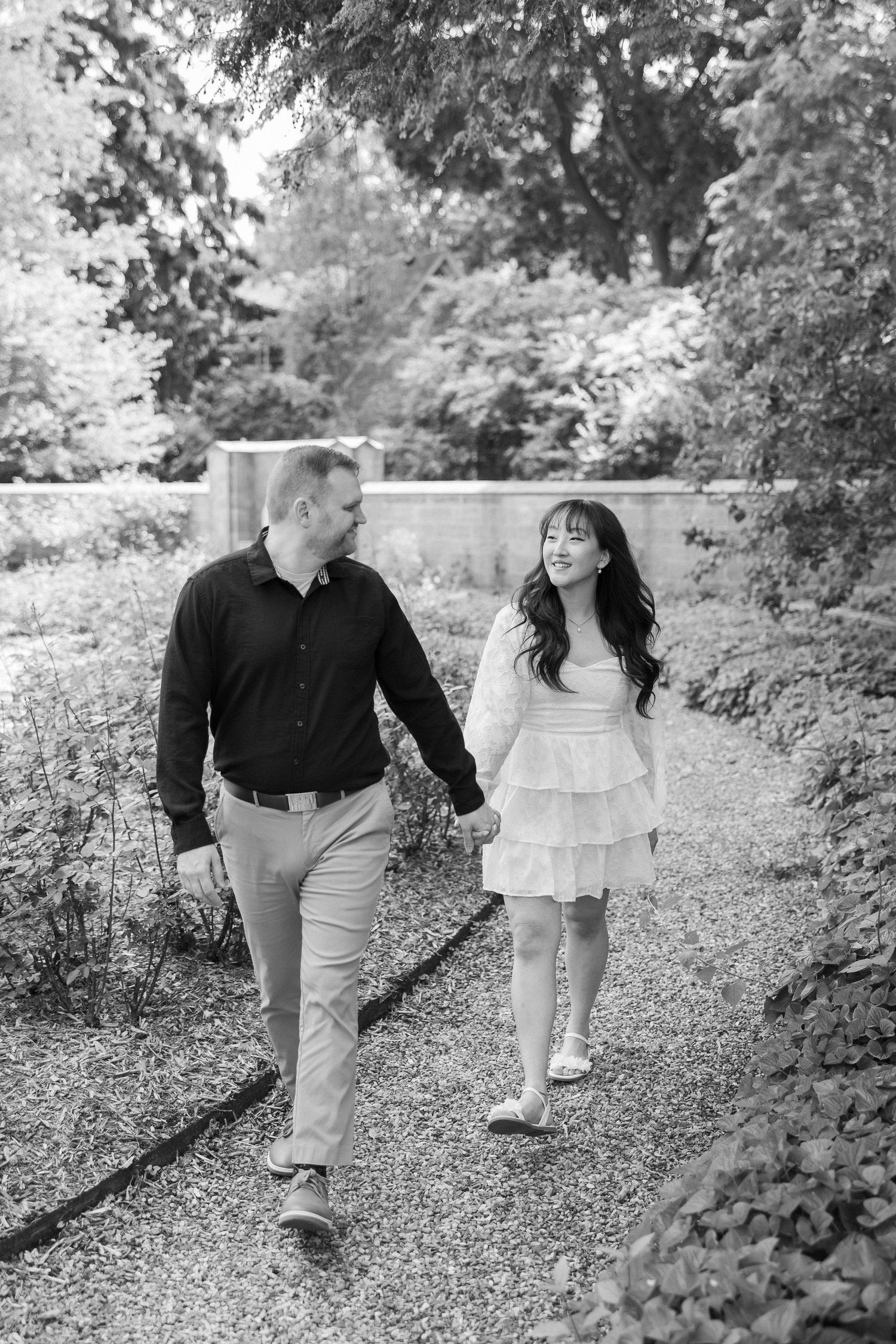 Couple takes romantic stroll on gravel path through park in black and white photography.