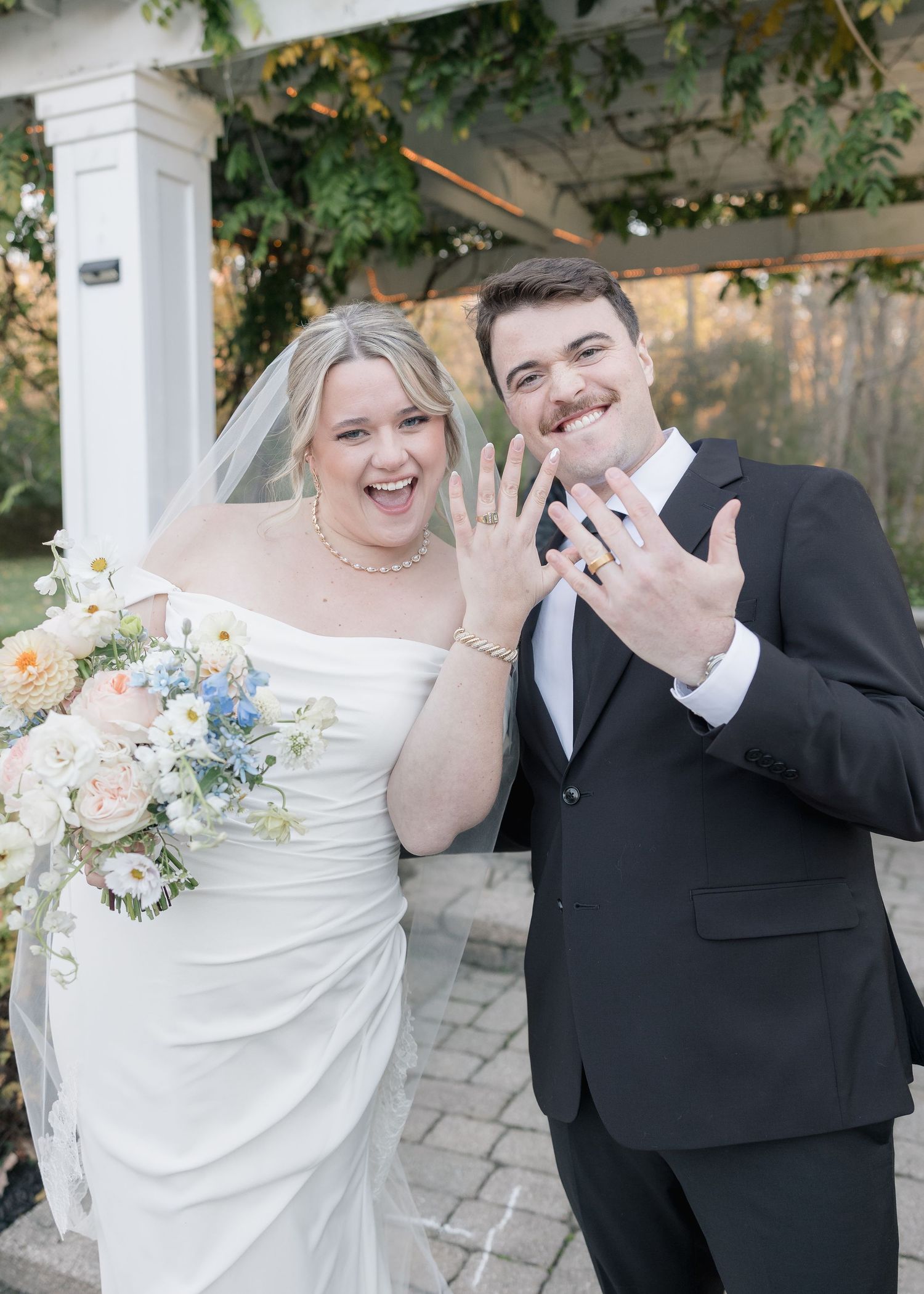 A newlywed couple showing off their wedding rings under a decorated pergola.