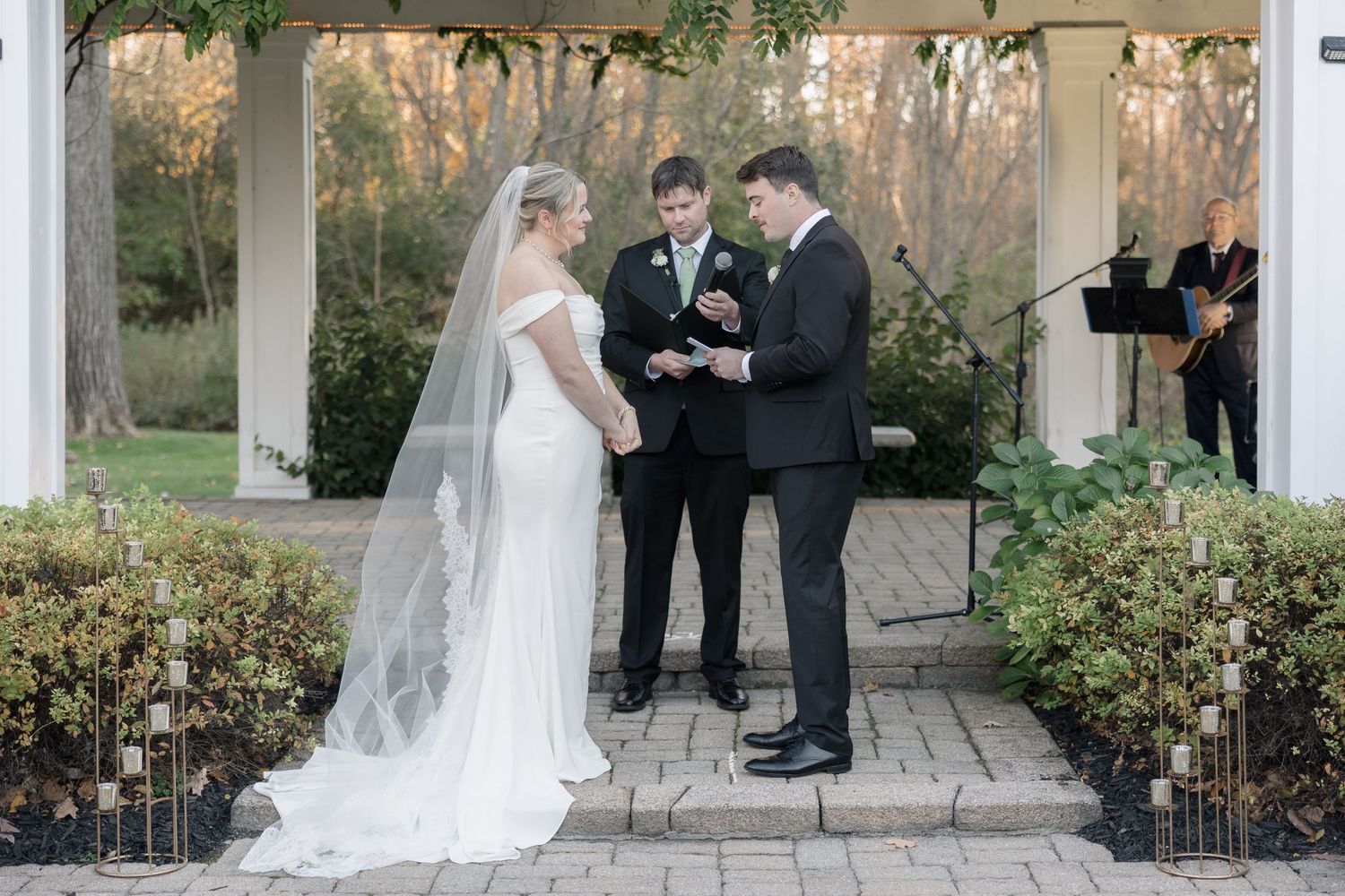 Wedding ceremony proceeds on stone patio under white pergola with natural greenery.