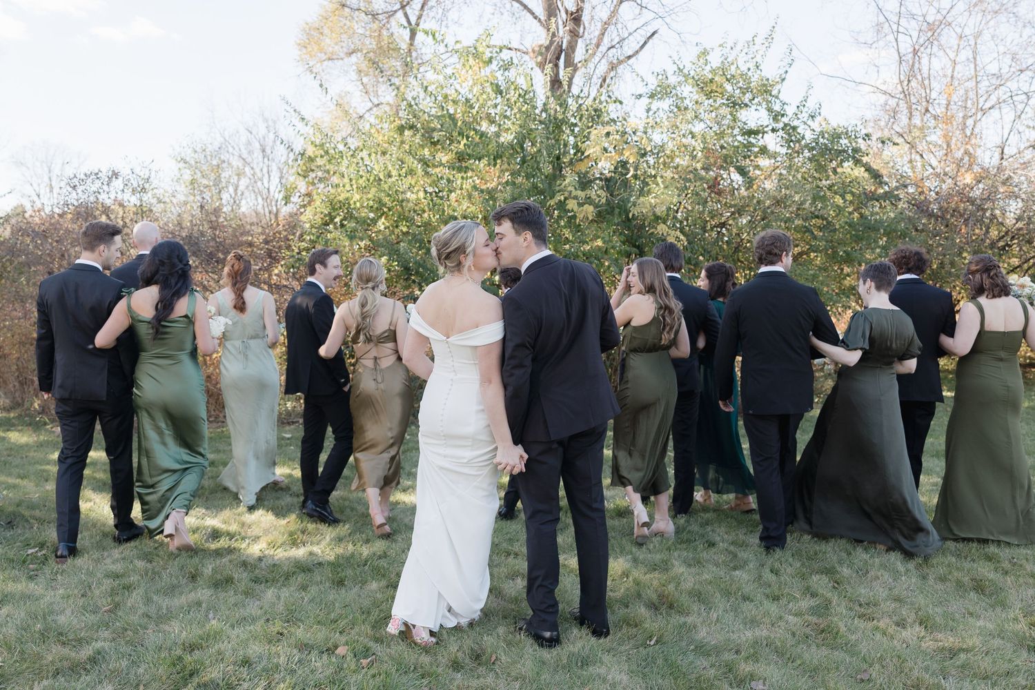 Wedding party in olive green dresses and black suits gather together on a grassy outdoor area during celebration.