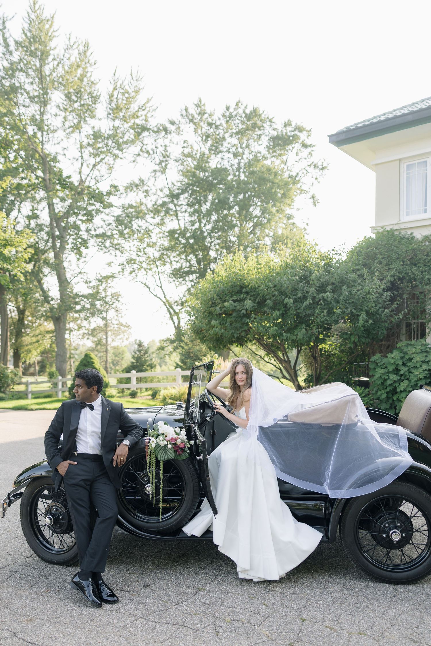 Bride and groom pose beside a vintage black car outdoors, surrounded by lush green trees on their wedding day.