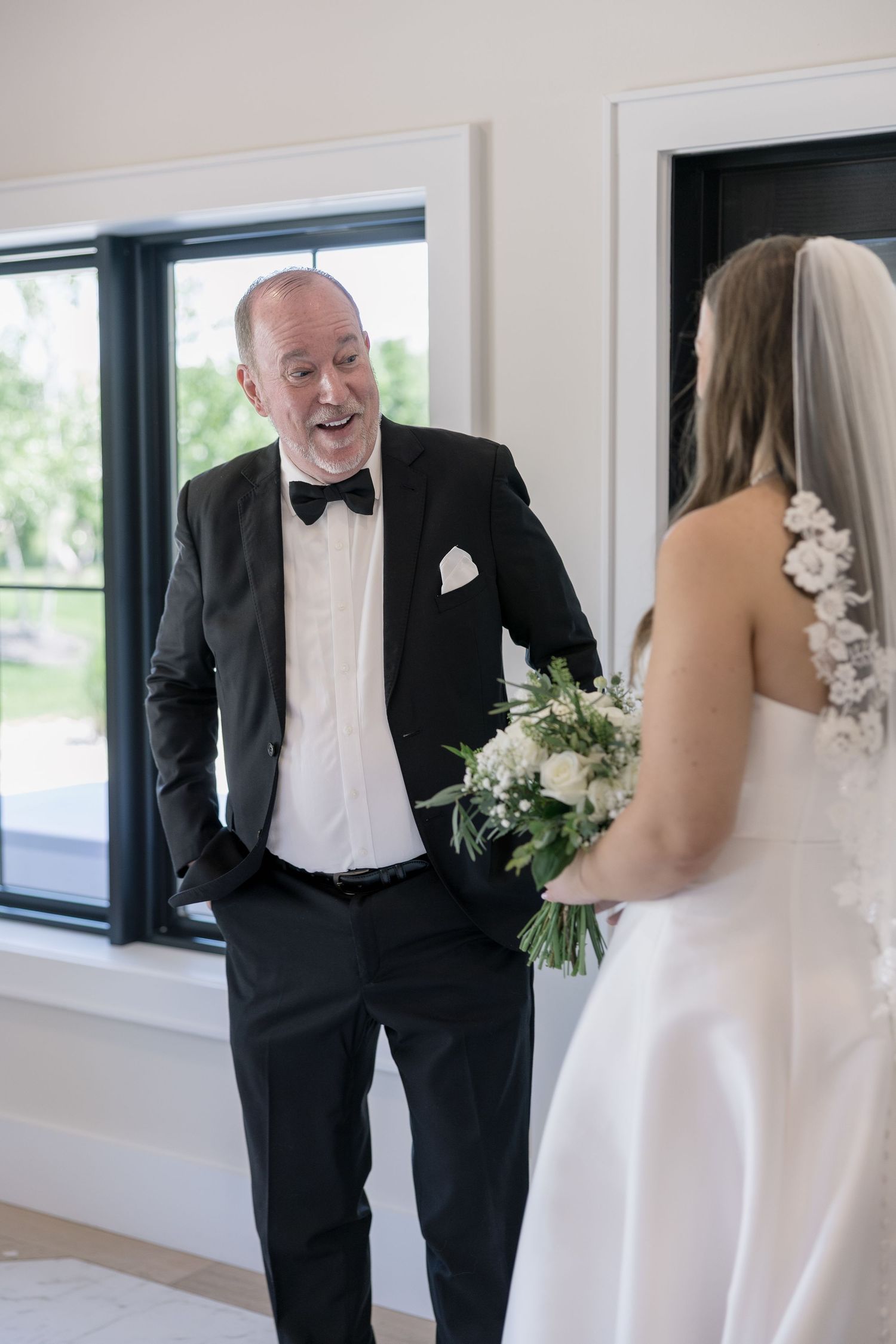 Father sees bride for first time, emotional first look moment with groom in tuxedo and bride holding bouquet.