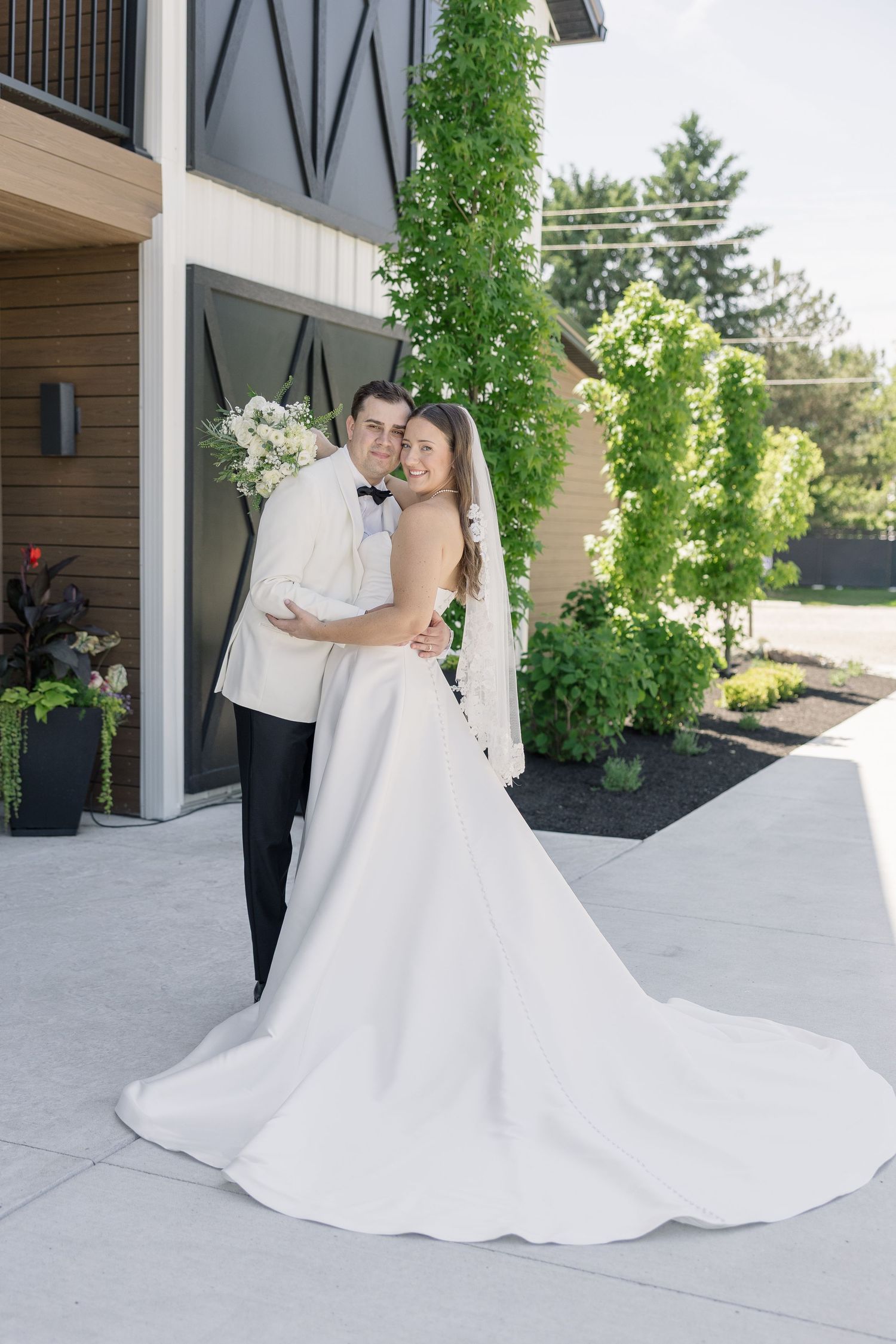 Bride in elegant white gown and groom in white tuxedo embracing outside modern wedding venue with greenery.