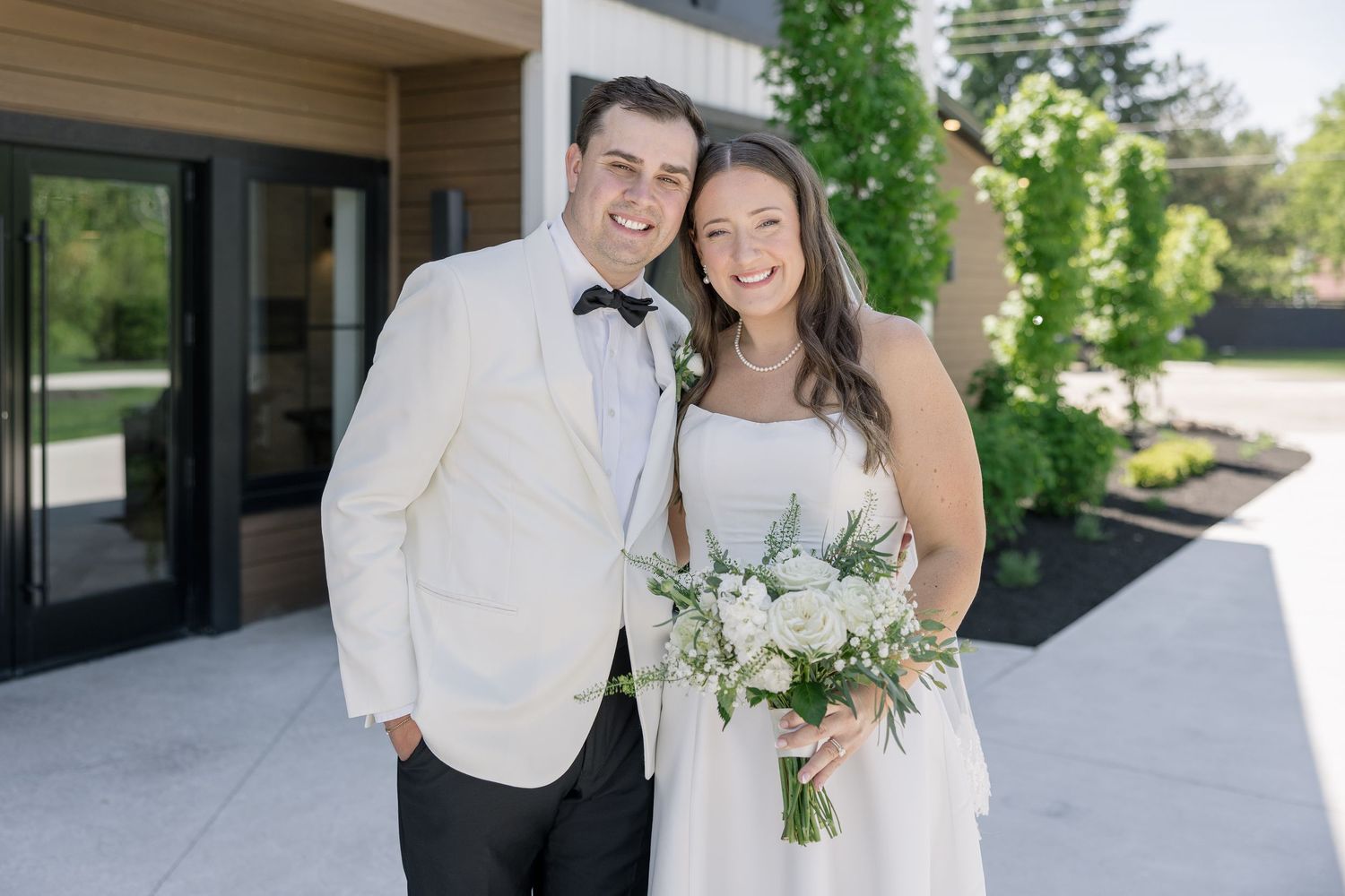 Smiling bride and groom pose outdoors, bride holding white floral bouquet, groom in white tuxedo jacket with bow tie.