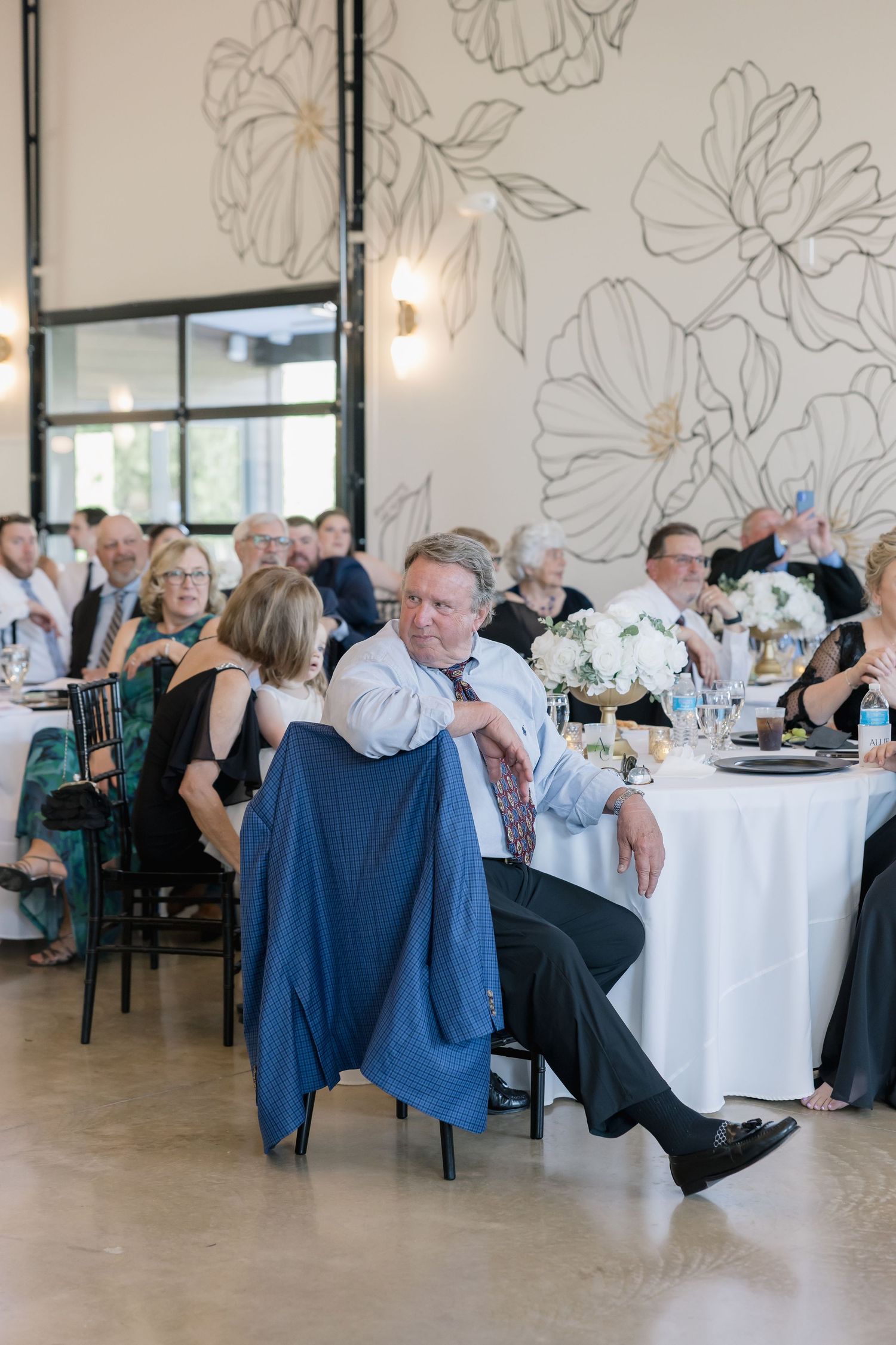 Wedding guests seated at decorated tables in an elegant venue with floral wall art.