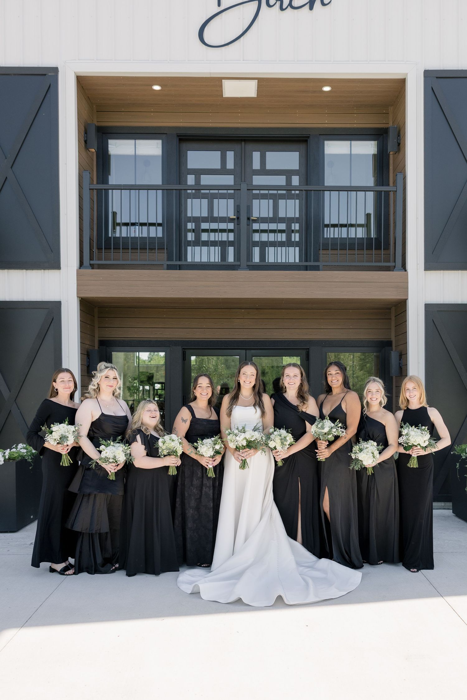 Bride in white gown poses with bridesmaids in black dresses holding white bouquets outside a modern farmhouse venue.