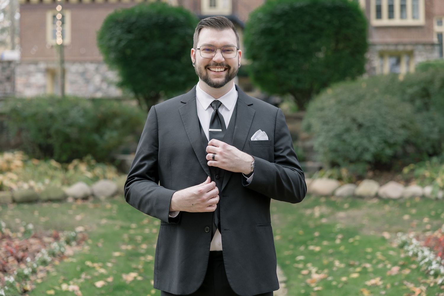 A person in a charcoal gray suit and tie smiles while standing in front of a landscaped garden and building.